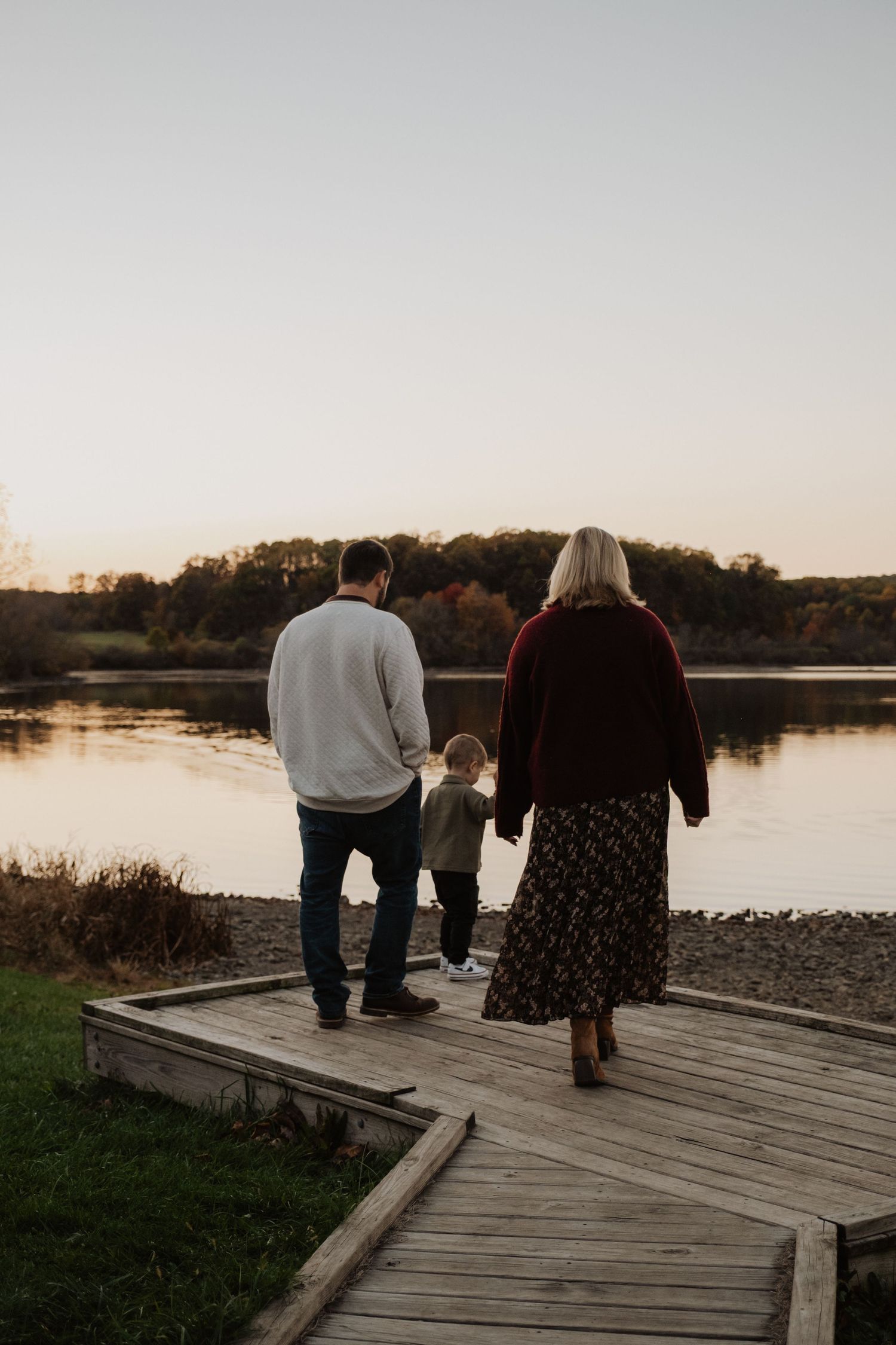 Family of three standing on wooden dock overlooking calm lake at sunset in autumn.