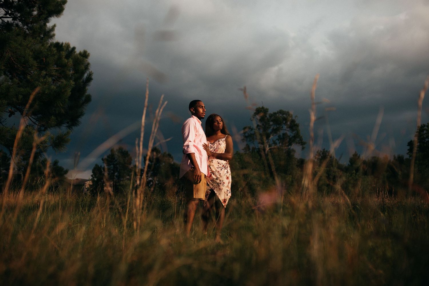 Couple embracing in golden field under dramatic stormy sky with rainbow at sunset.