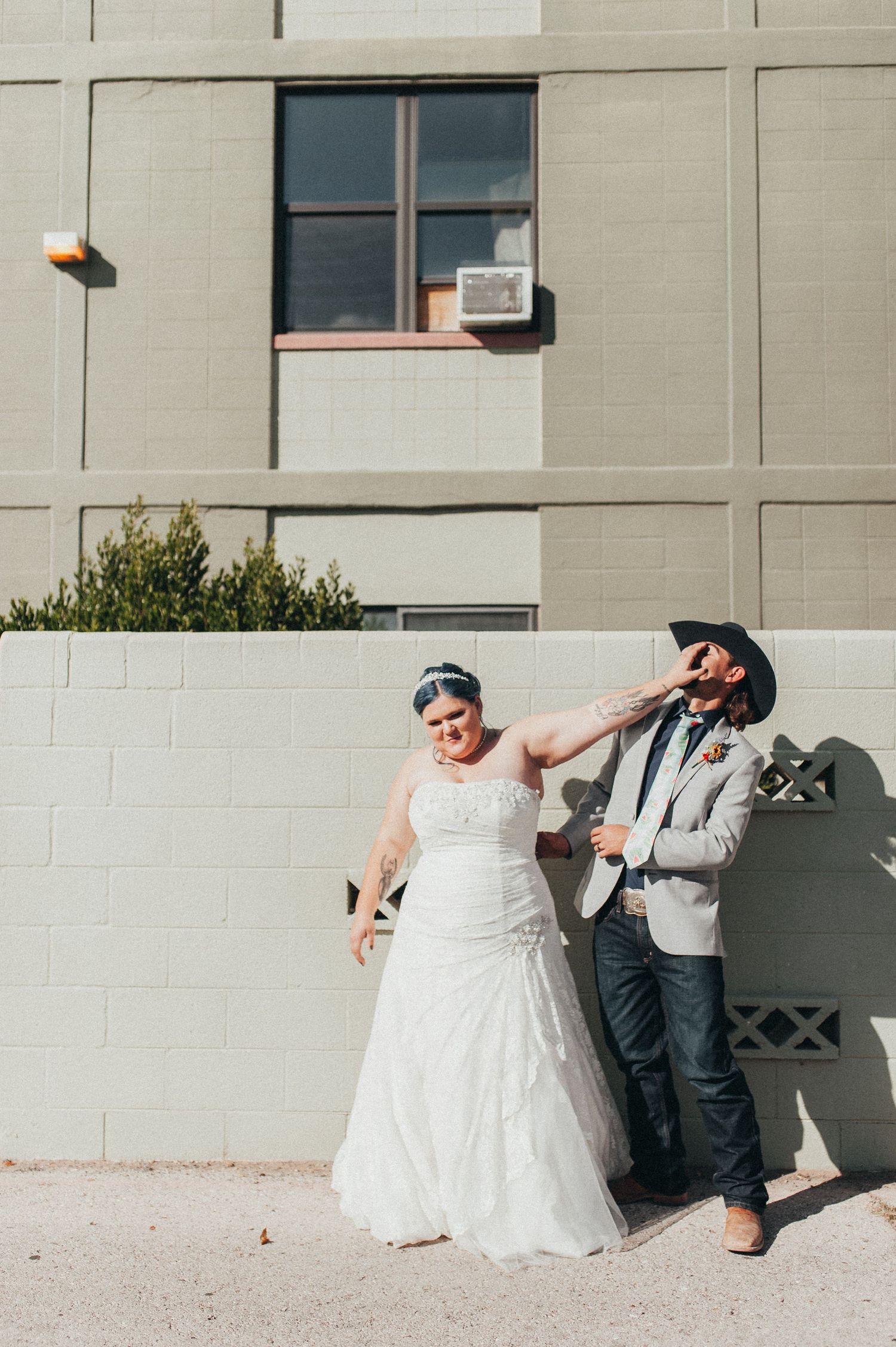 Tattooed bride and groom in cowboy hat pose playfully outside brick building on their wedding day.