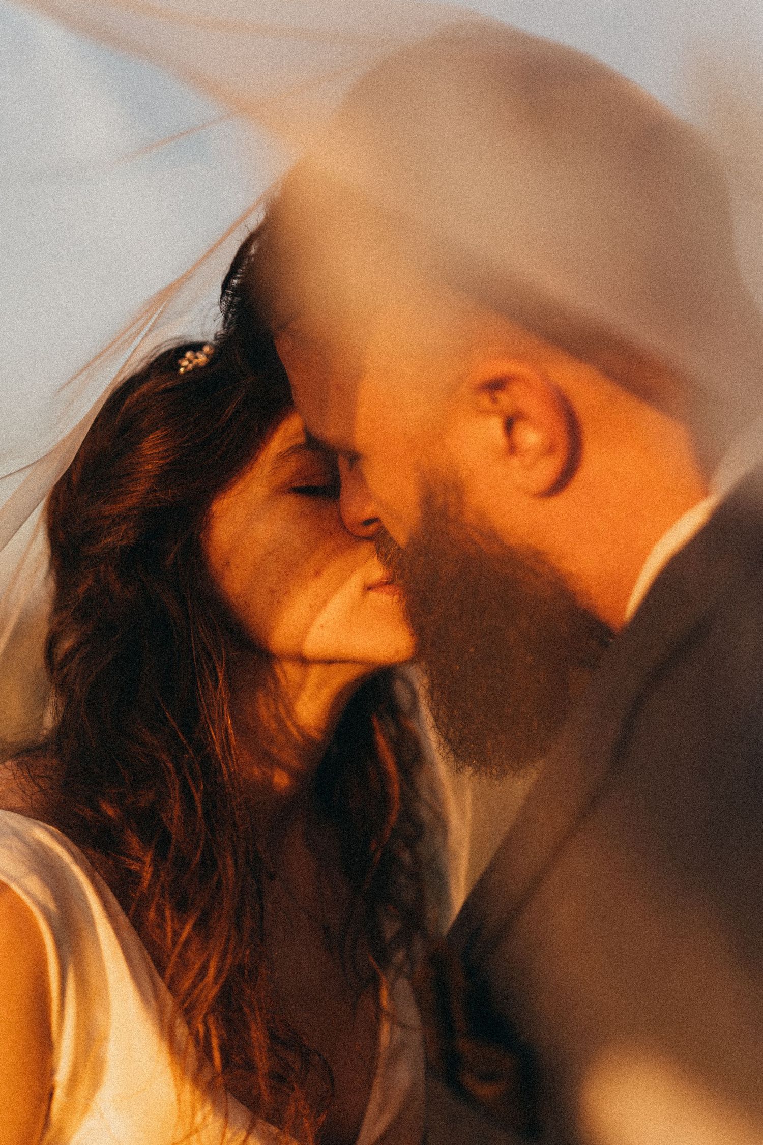 Romantic golden hour wedding photo of bride and groom sharing a kiss under a flowing veil.