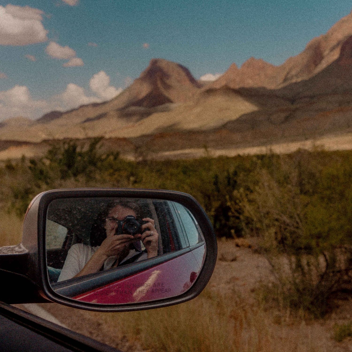 Person taking a selfie in a car side mirror with scenic desert mountains in the background.