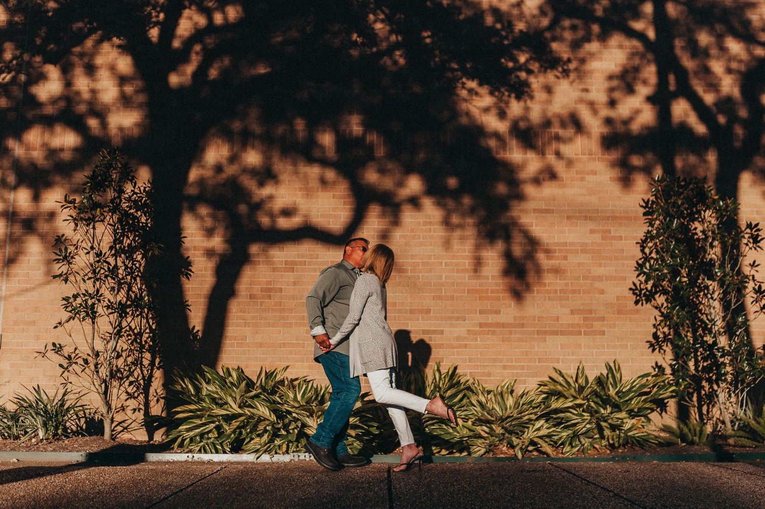Couple sharing a romantic kiss against a sunlit brick wall with dramatic tree shadows in an outdoor setting.