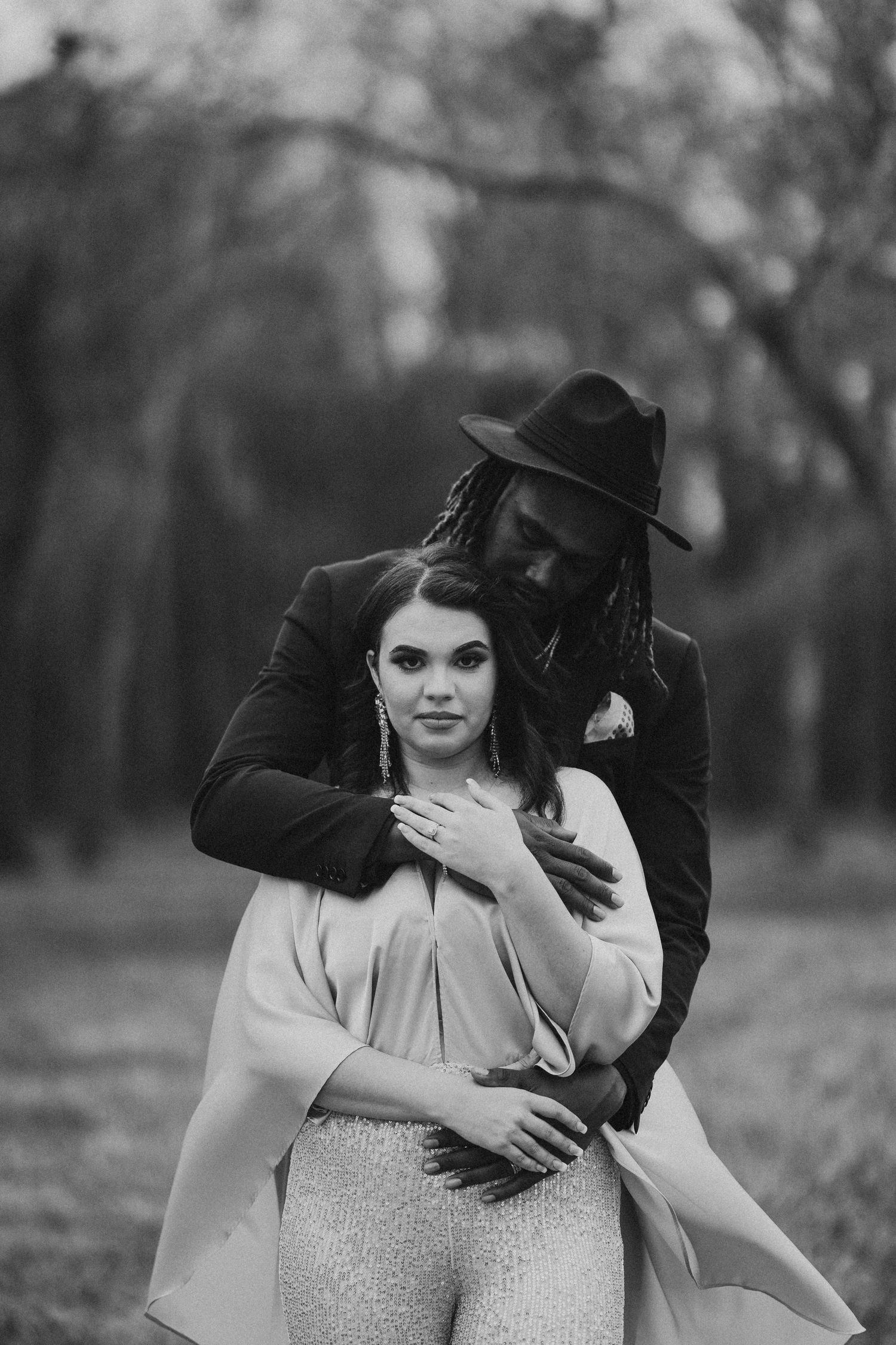 Black and white photo of an interracial couple embracing outdoors, man in hat with dreadlocks hugging woman in elegant attire.