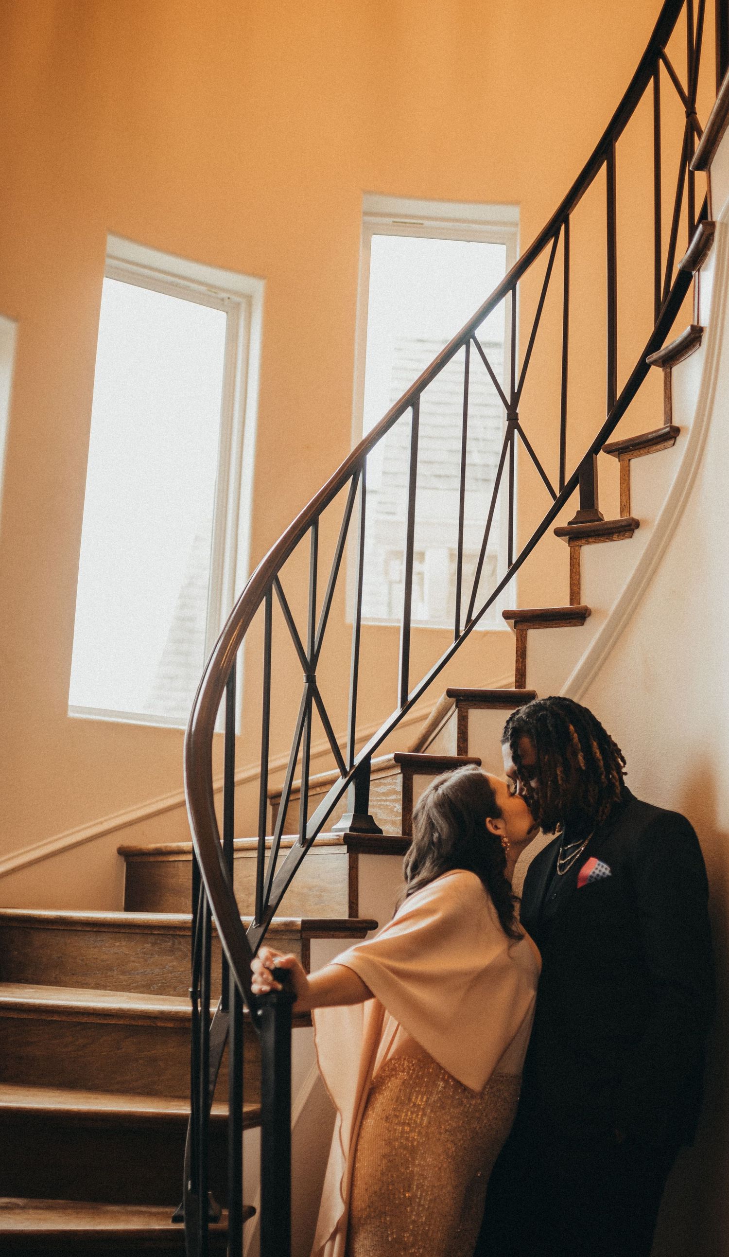 Romantic couple sharing a kiss on an elegant spiral staircase in a warmly lit home.