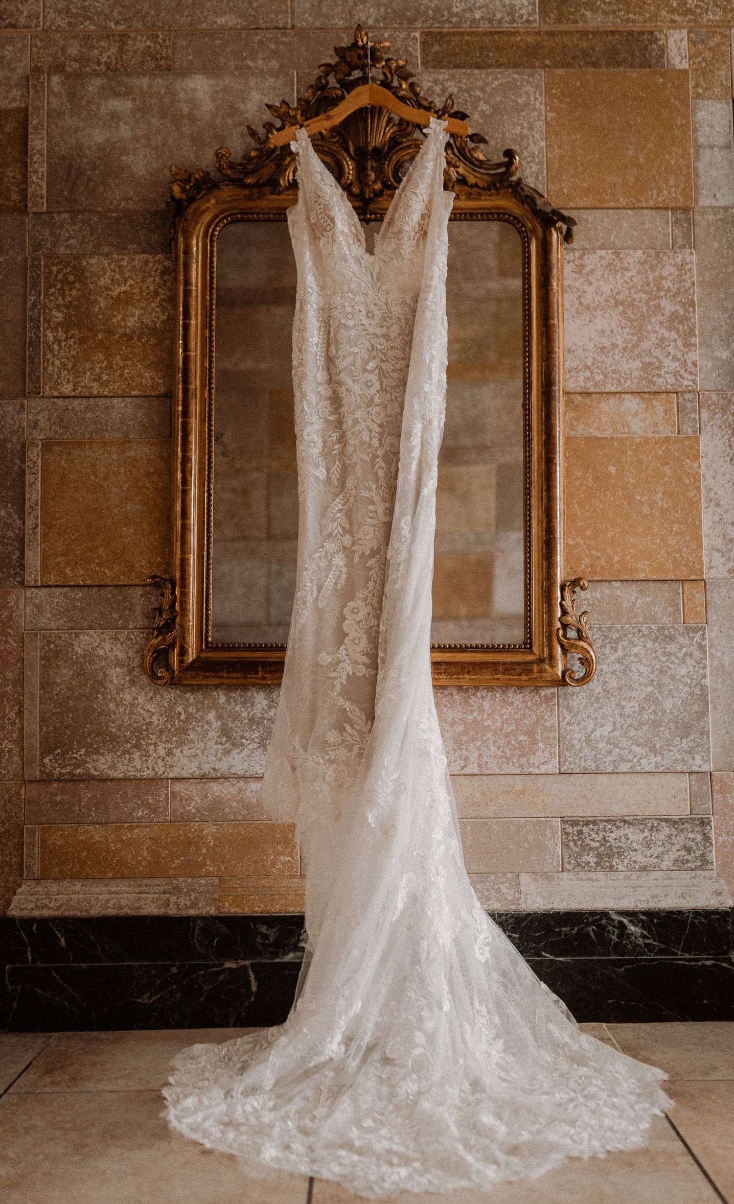 Elegant lace wedding dress with train hanging on ornate gold mirror against stone wall.