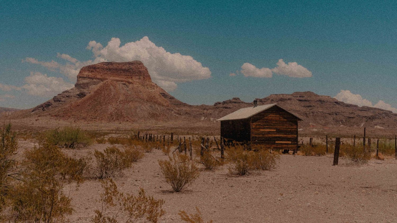 Rustic wooden shack in arid desert landscape with dramatic mesa formations under blue sky.
