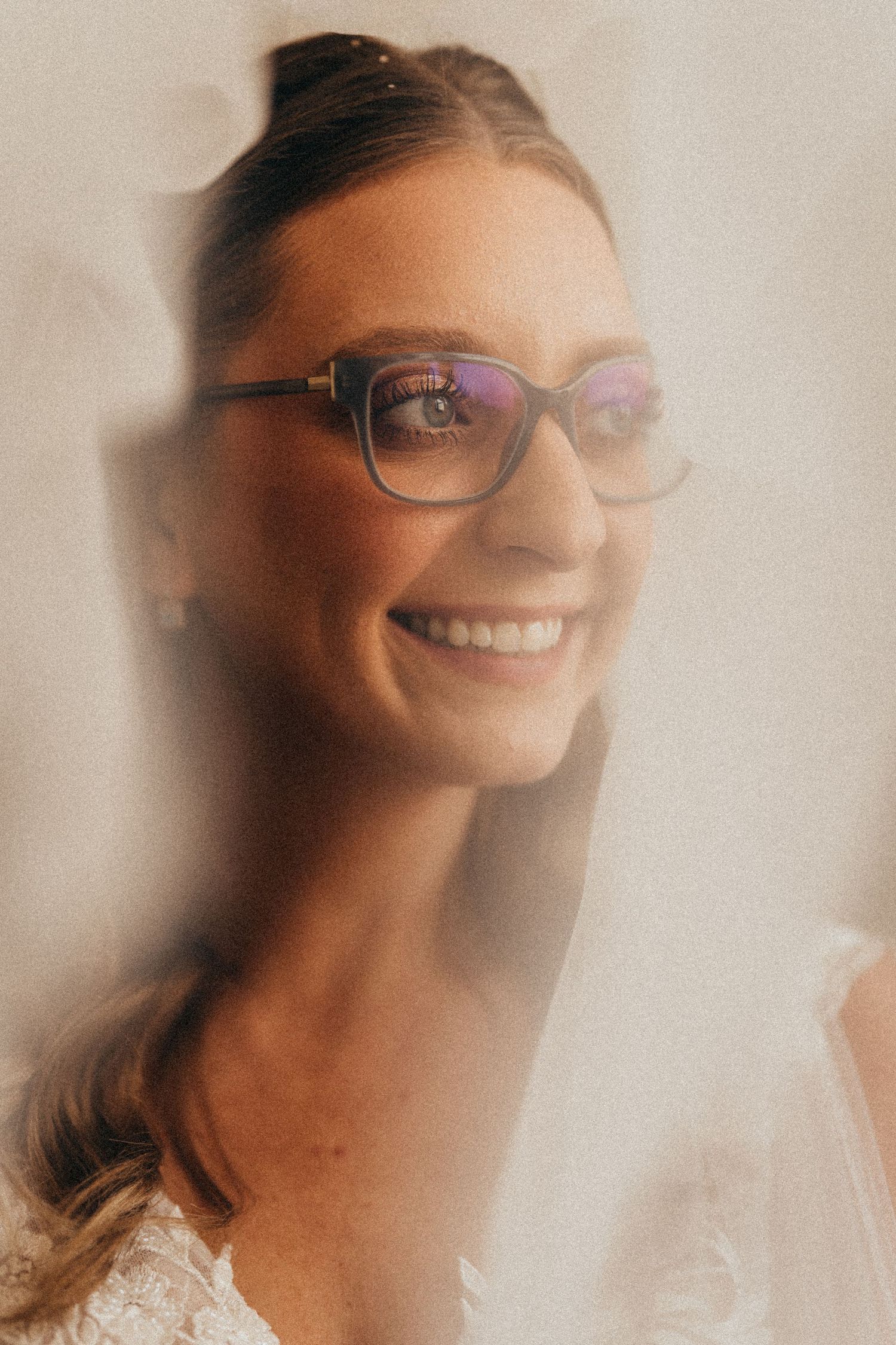 Smiling bride in glasses and lace dress, photographed through a sheer veil in a soft, dreamy portrait.