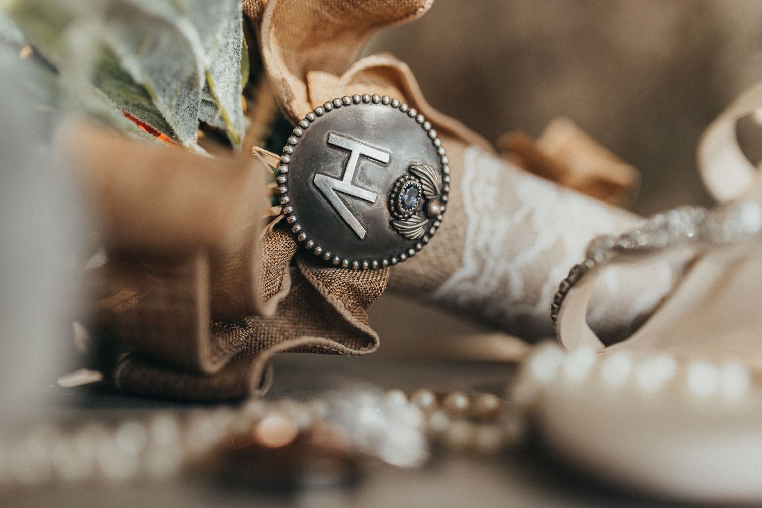 Close-up of a decorative silver monogram brooch with floral detail beside cowboy boots and pearl jewelry.