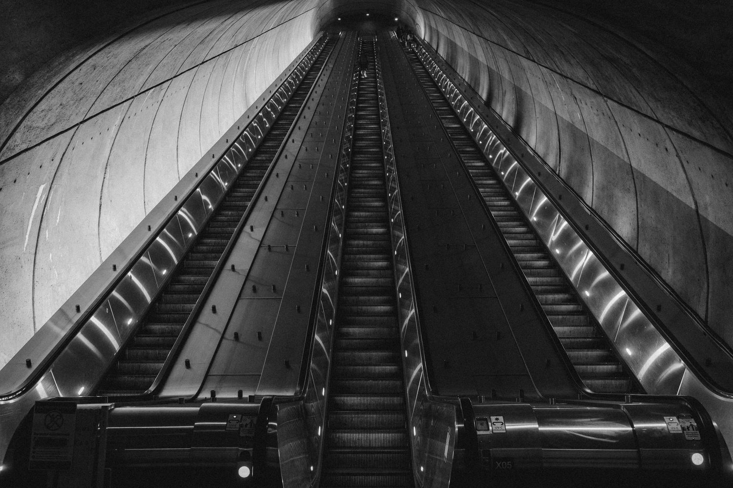 Black and white symmetrical view looking up a long escalator tunnel in a subway station.