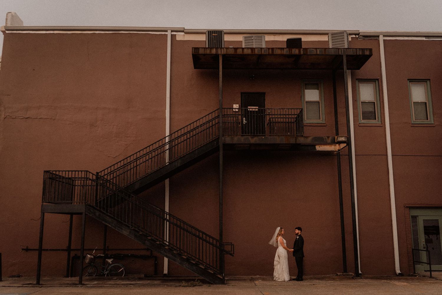 Couple shares a romantic moment beneath an iron fire escape staircase on the exterior of a rustic brick building.