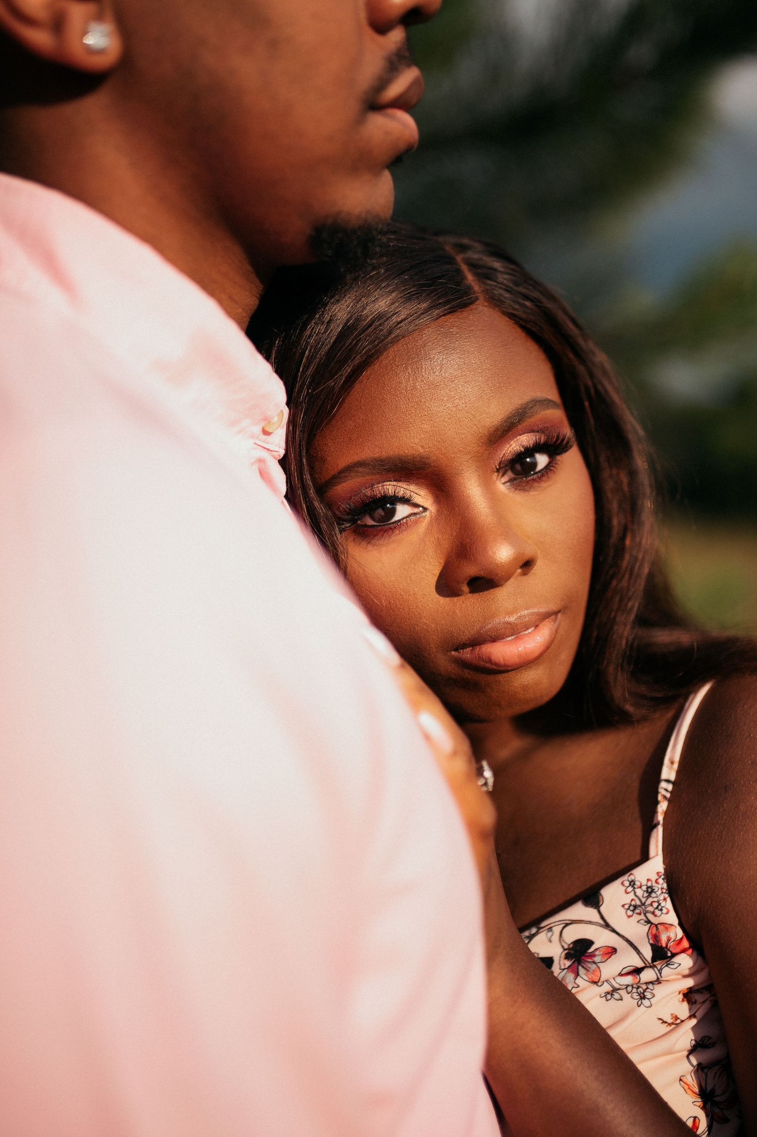 A young Black woman in a floral dress gazes thoughtfully while standing close to a man in a pink shirt outdoors.