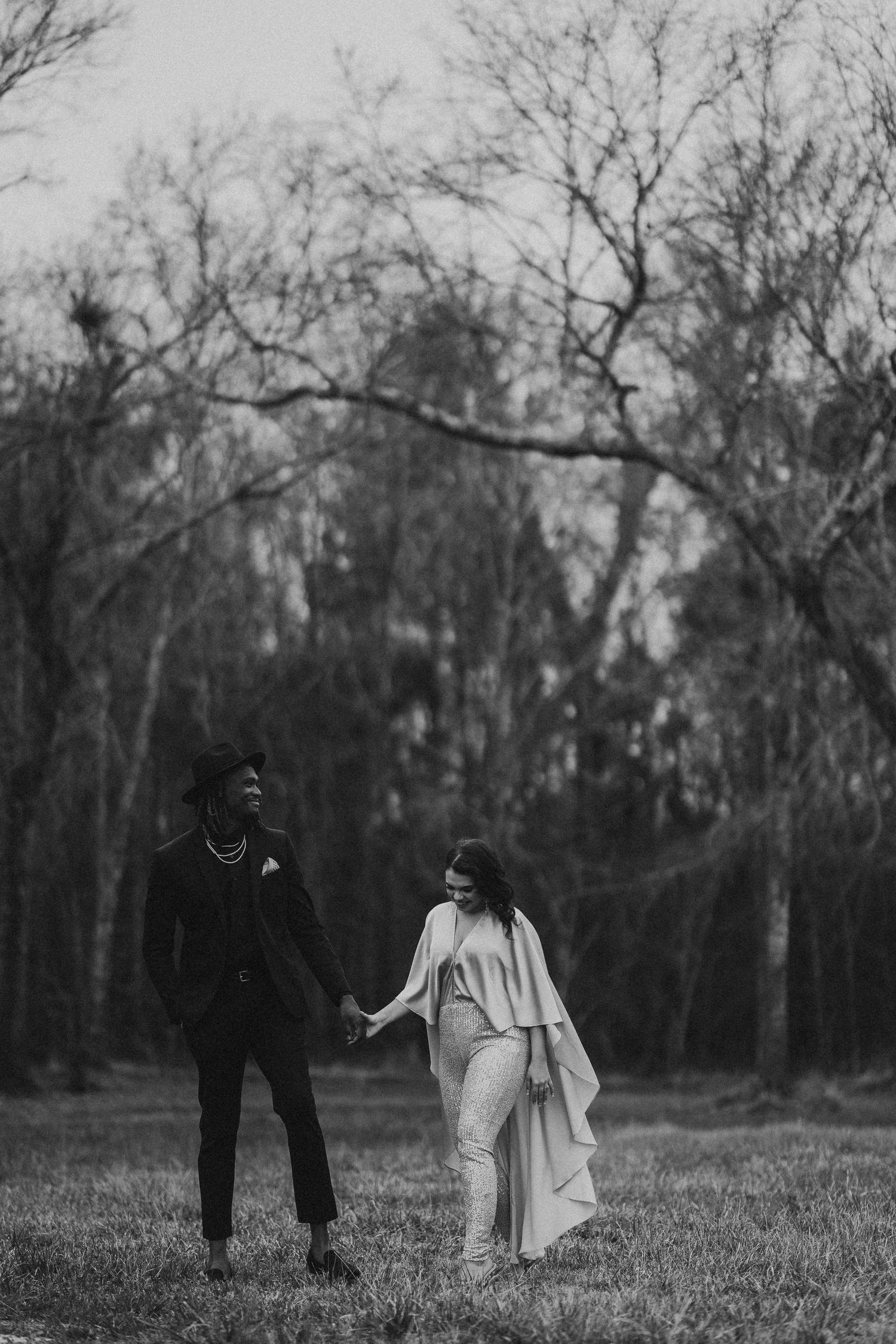 Black and white photo of a couple holding hands in a field surrounded by bare trees.
