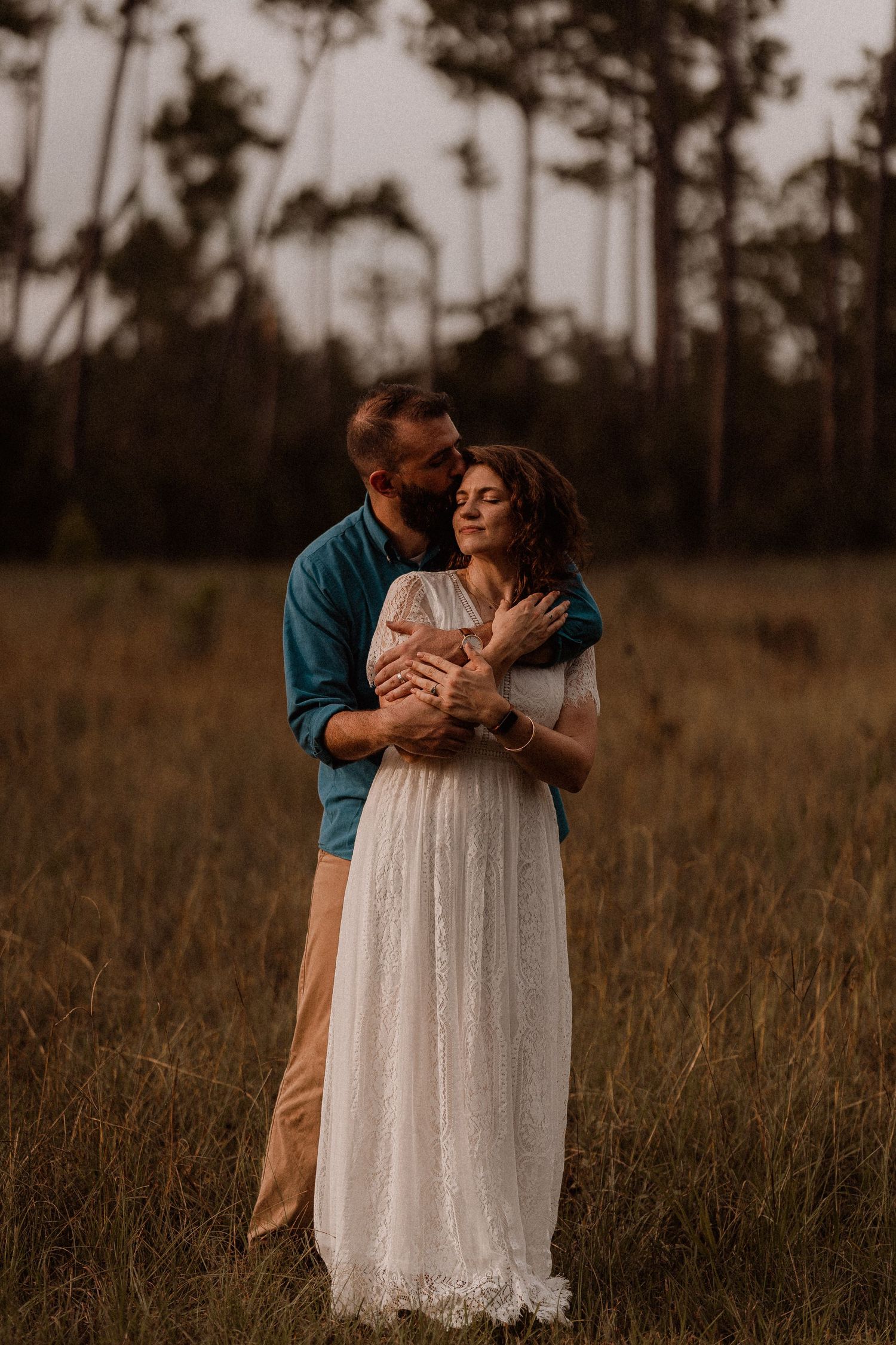 Couple embracing in a golden field at dusk, woman in white lace gown, man in teal shirt kissing her head.