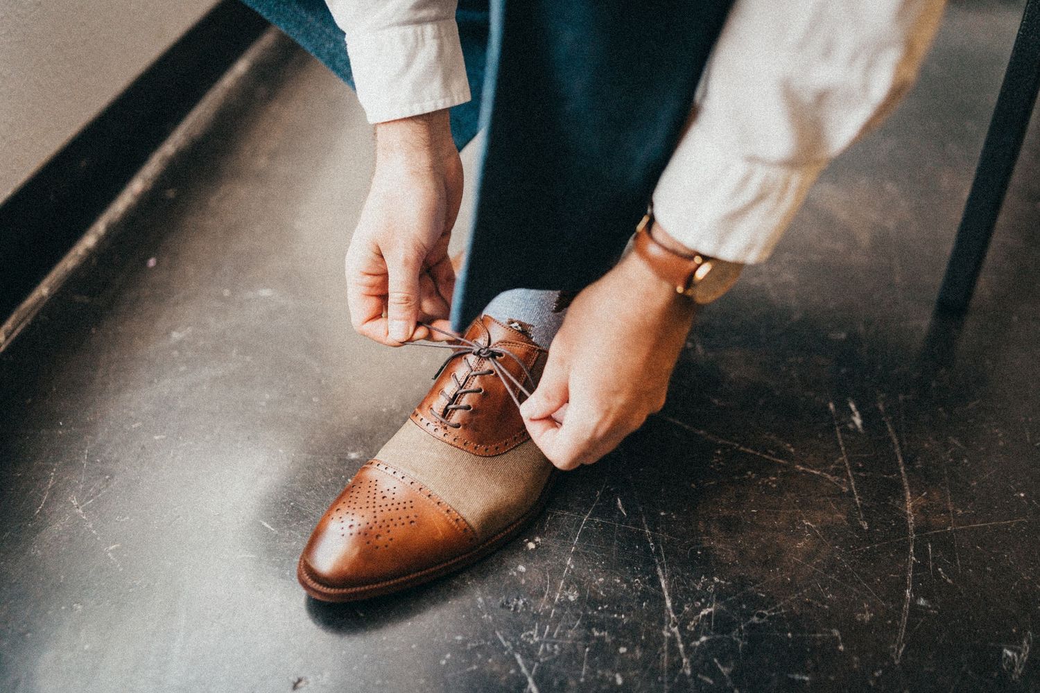Person tying brown leather brogue oxford dress shoes while wearing a gold watch and white trousers.