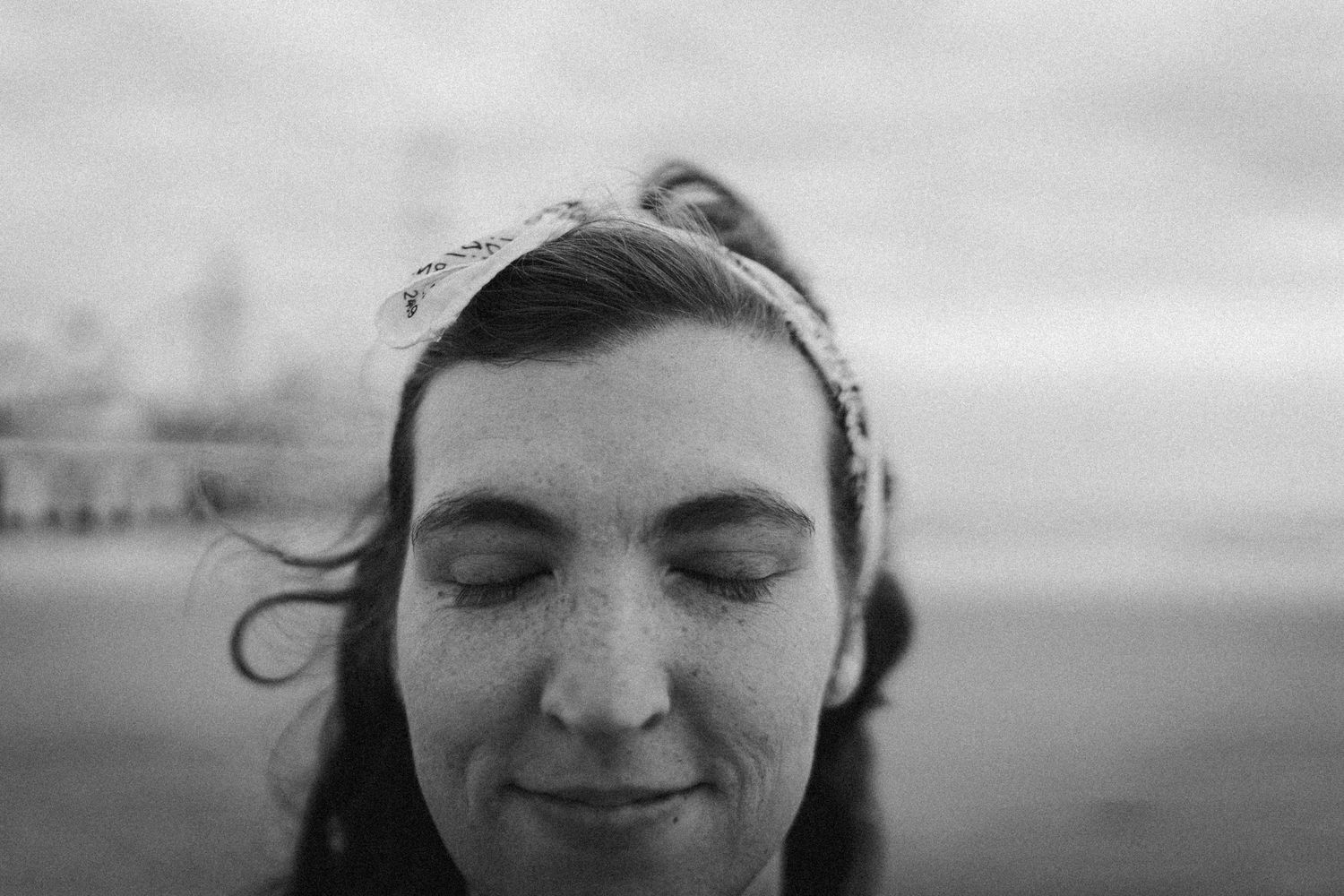 Black and white portrait of a young woman with freckles, closed eyes, and a headband outdoors near water.