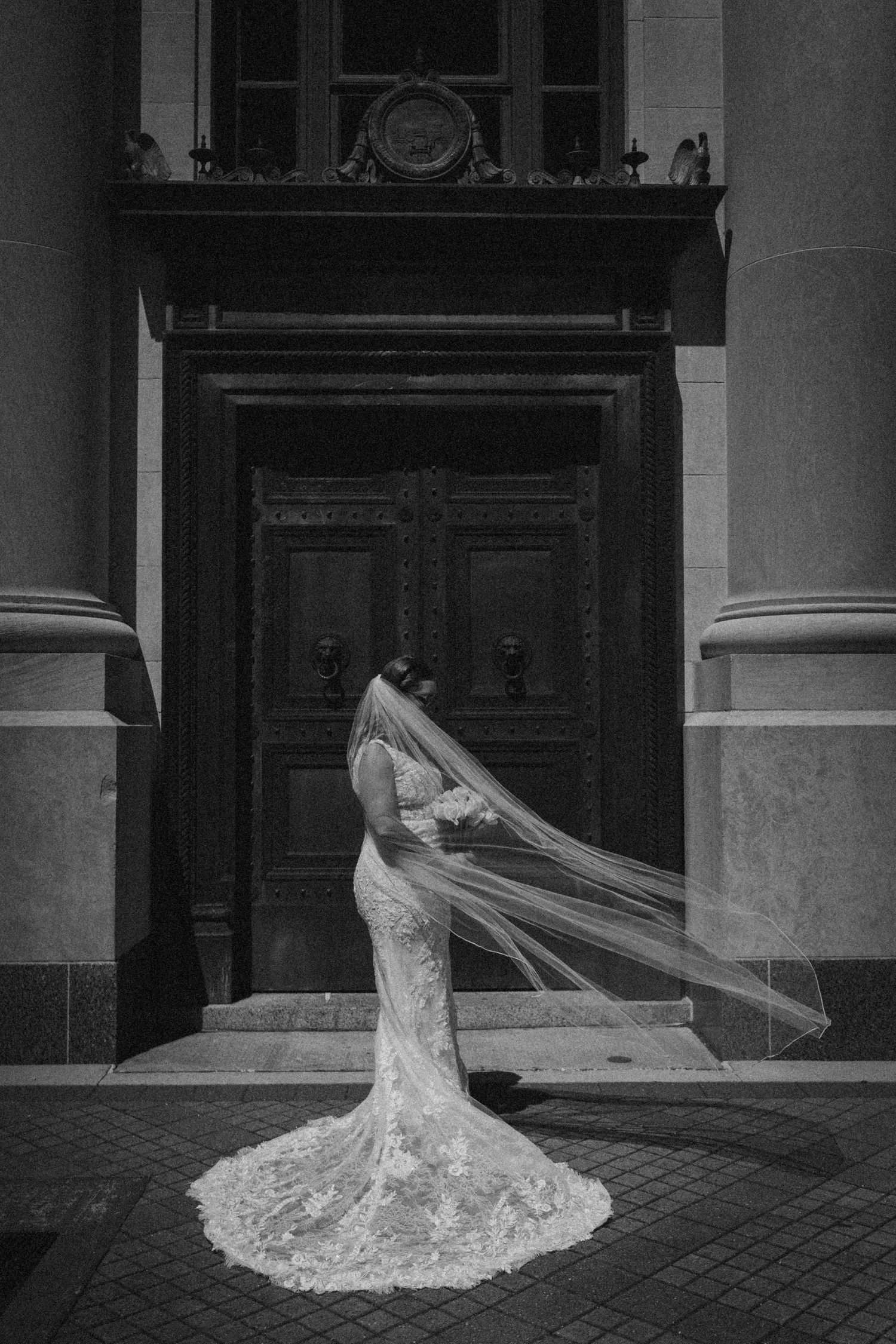 Black and white photo of a bride in lace gown with flowing veil standing before ornate doors.