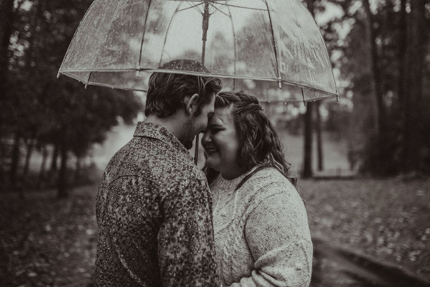 Couple sharing a romantic moment under a clear umbrella in the rain, in a black and white photo.
