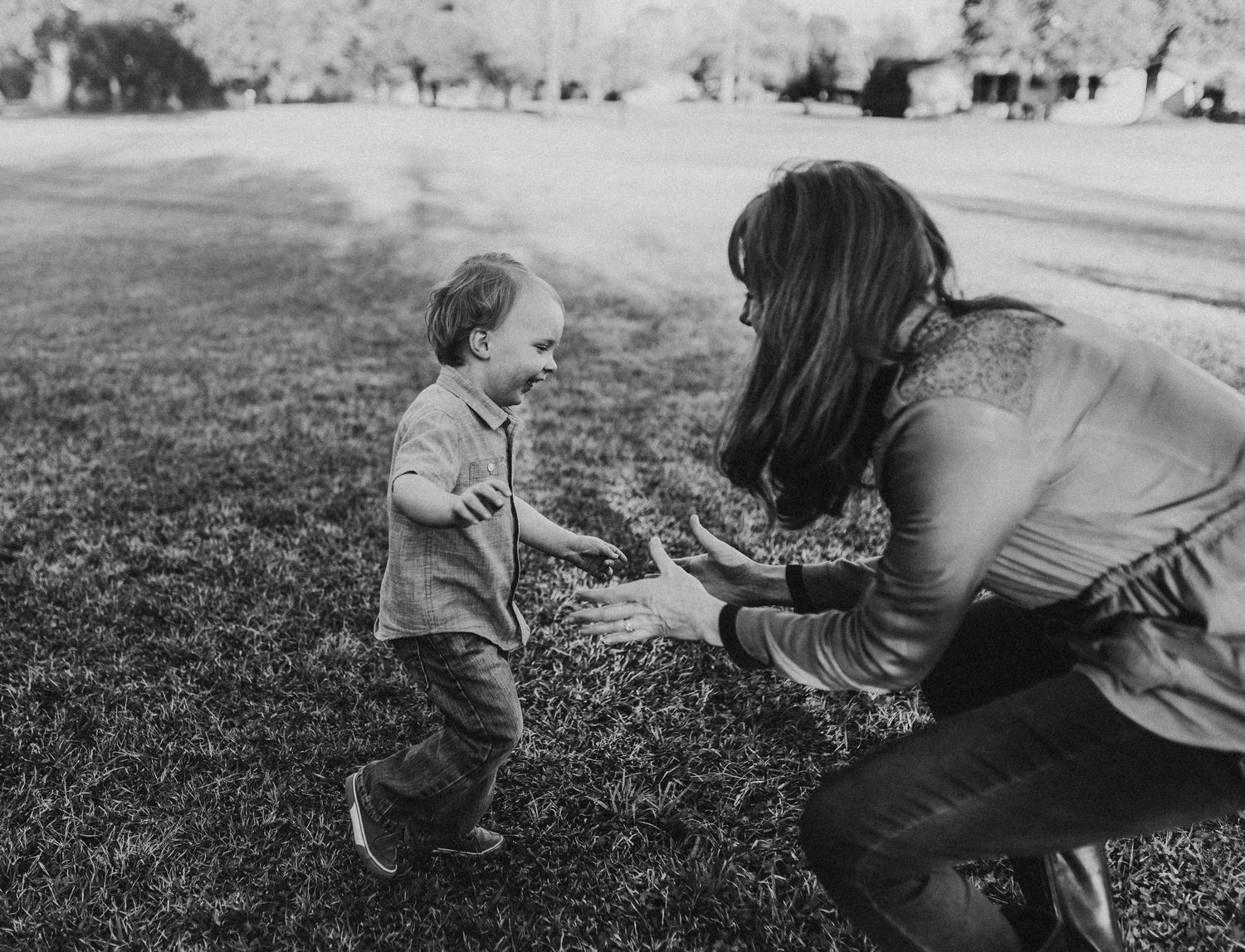Black and white photo of a toddler running toward a woman crouching with open arms on a grassy lawn.