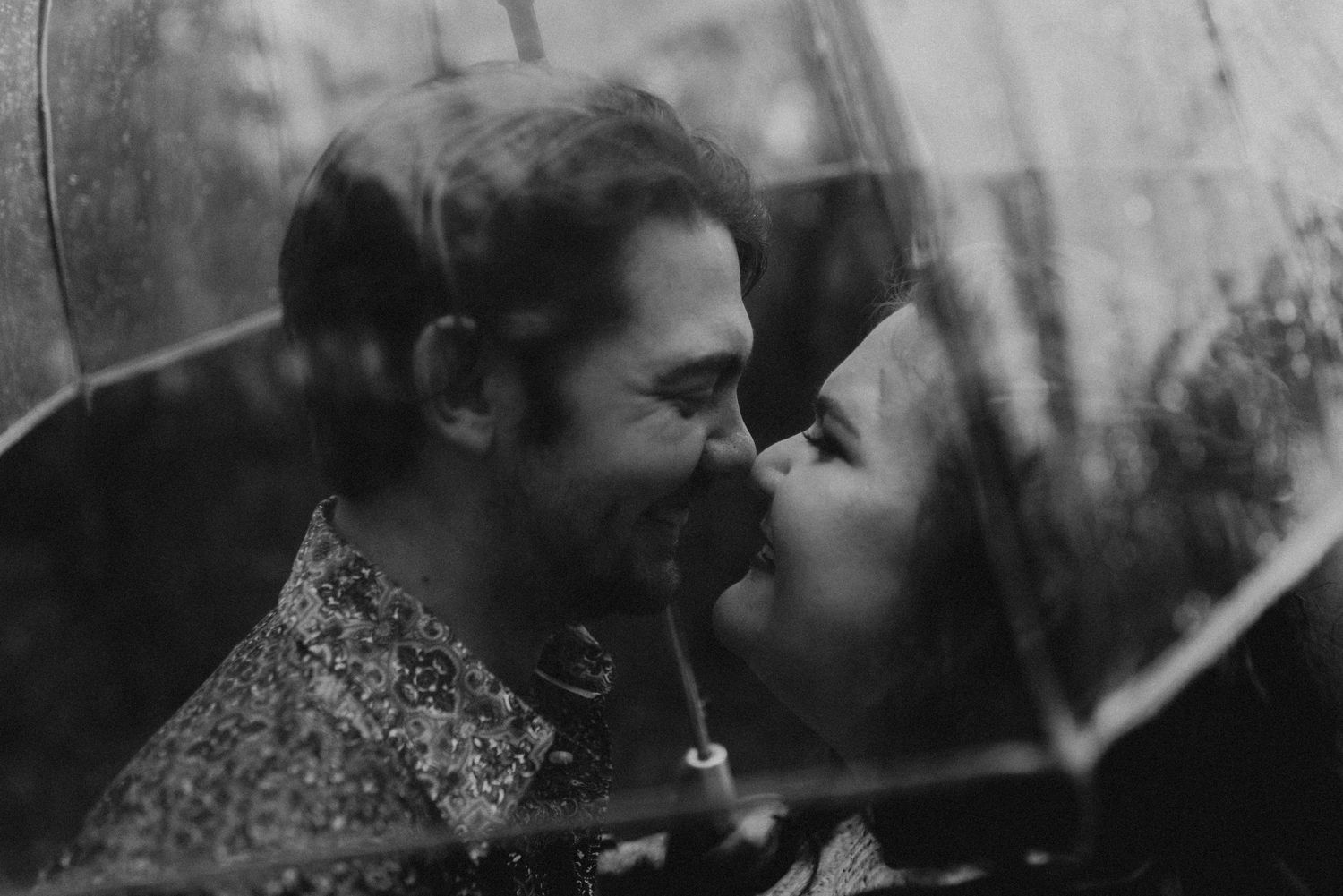 Black and white photo of a couple sharing a romantic moment under a clear umbrella in the rain.