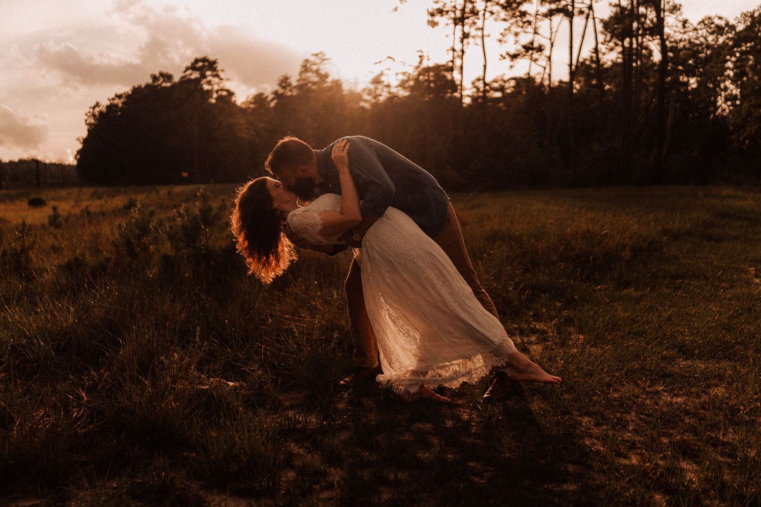 Couple sharing a romantic dip kiss at golden sunset in a lush green meadow with trees in background.