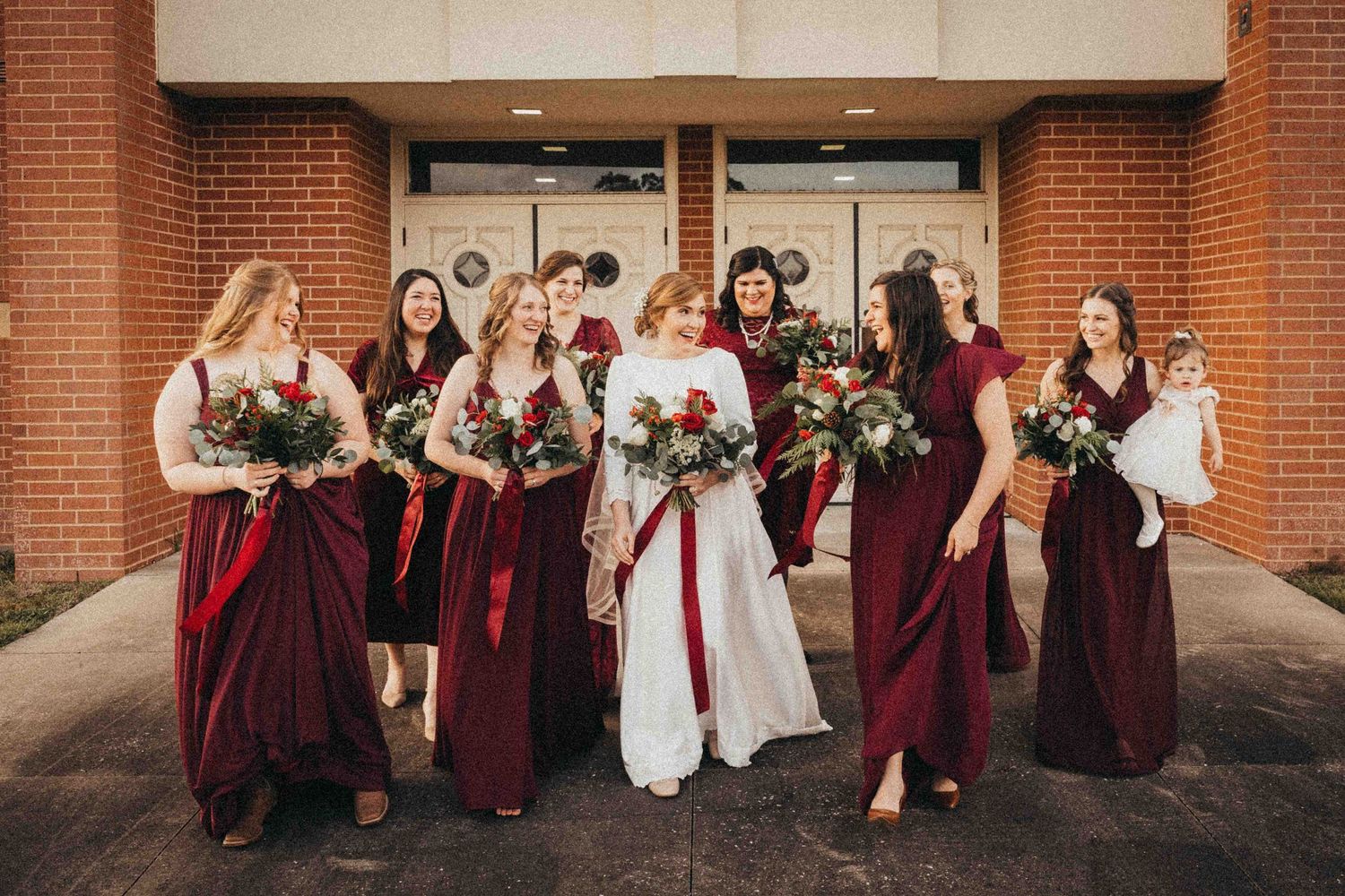 Bride in white gown walks with bridesmaids in burgundy dresses holding red floral bouquets outside brick building.
