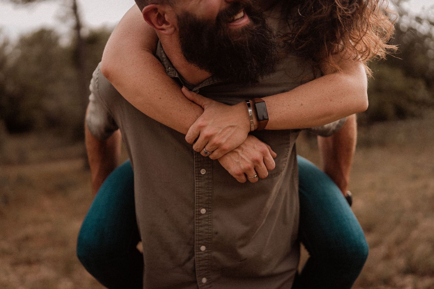 Bearded man giving a woman a piggyback ride outdoors in a field, both laughing and embracing joyfully.