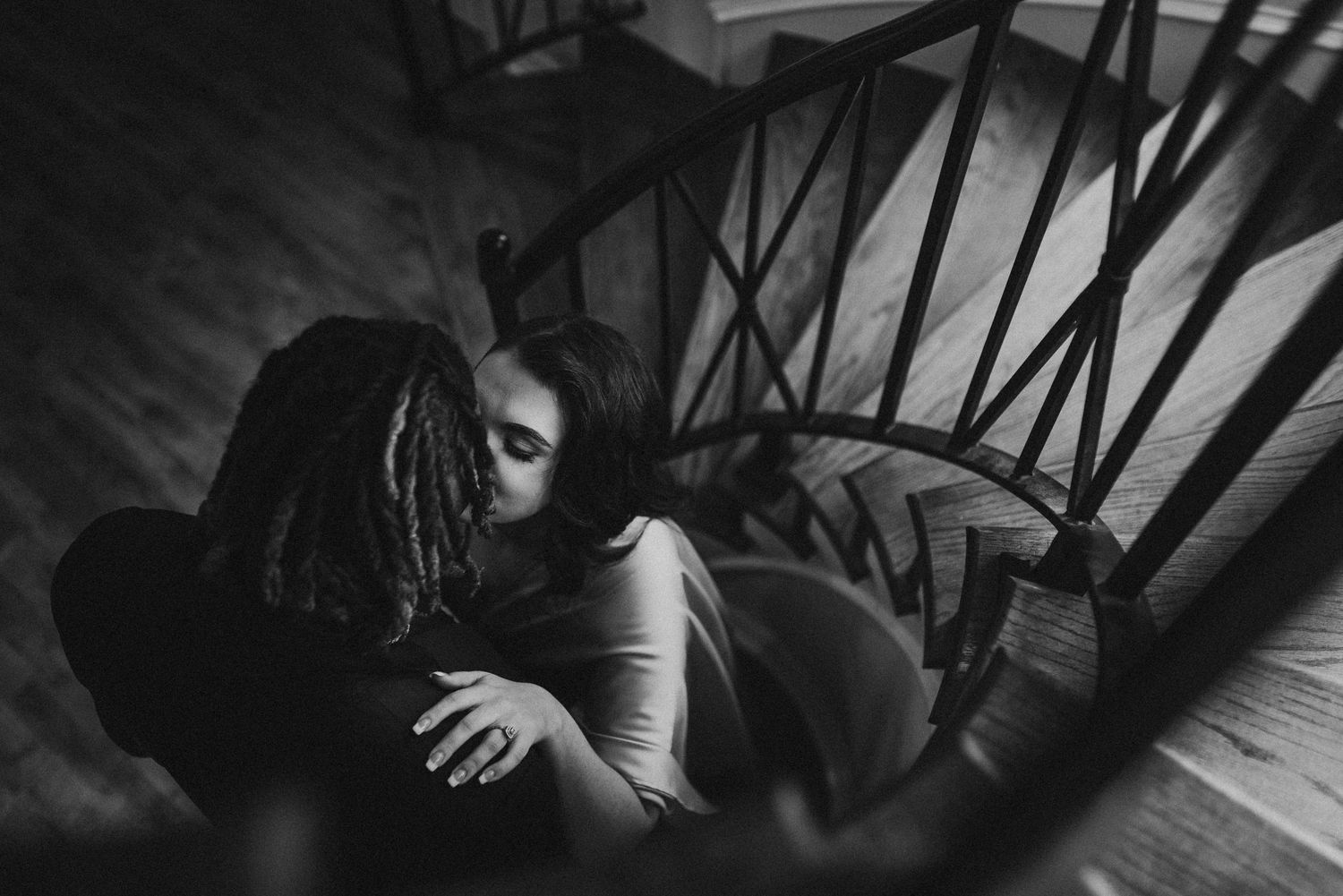 Black and white photo of a couple embracing on a spiral staircase in a romantic moment.