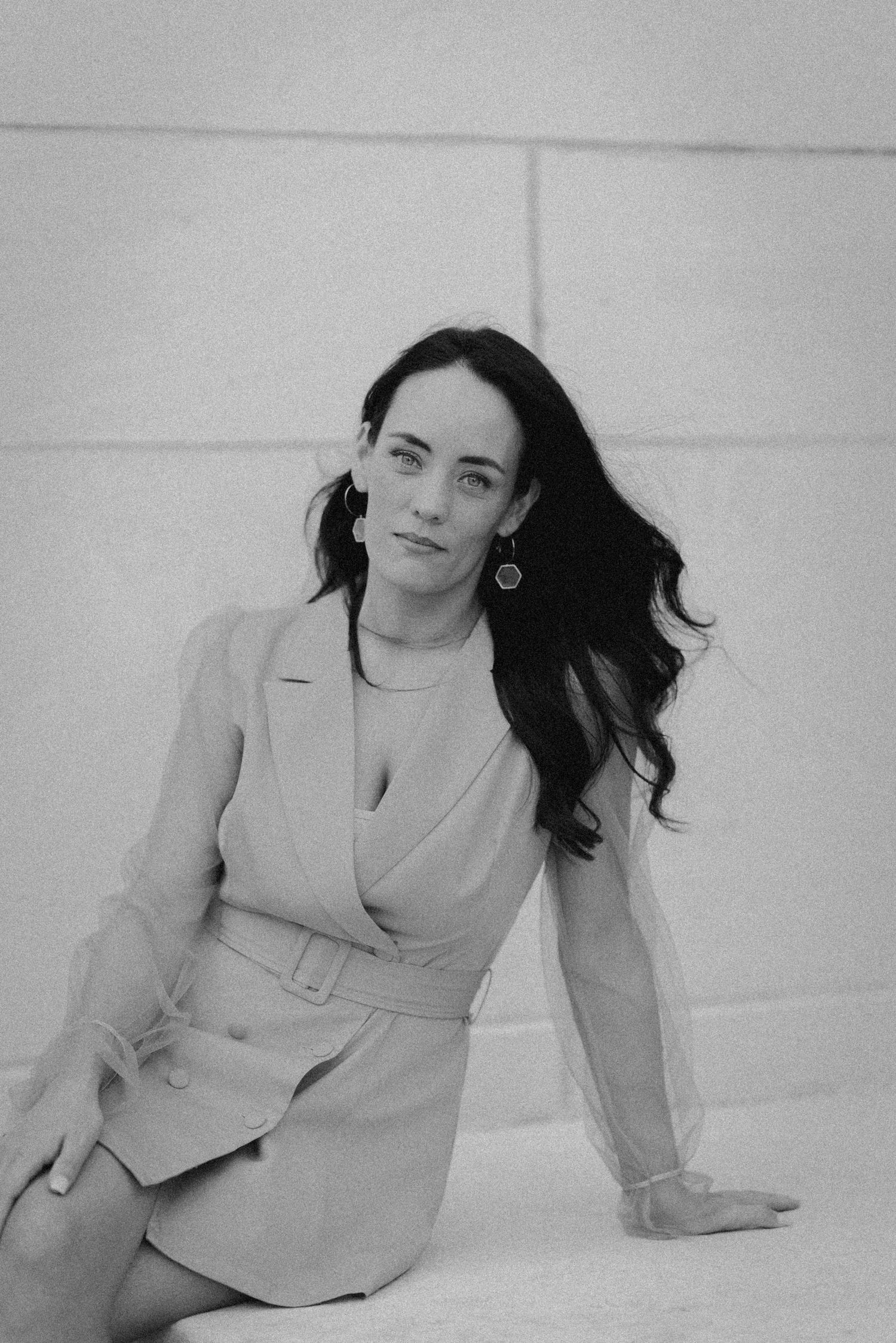Black and white portrait of a woman in a belted blazer dress with dark wavy hair and statement earrings.