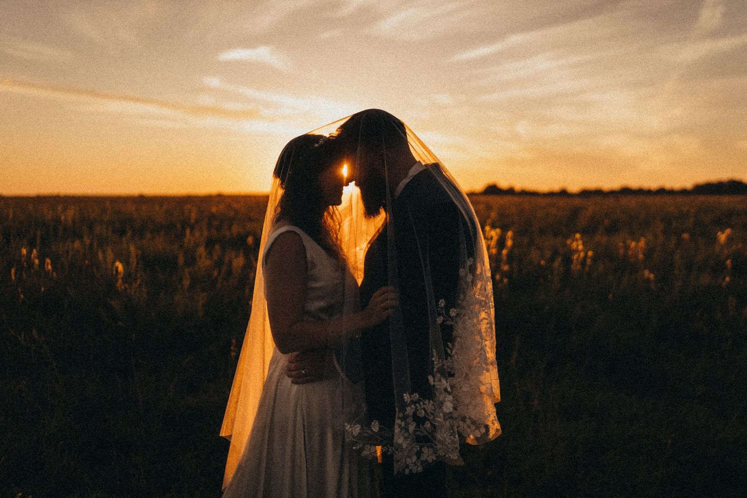 Bride and groom sharing a romantic moment under a wedding veil at golden sunset in a scenic field.