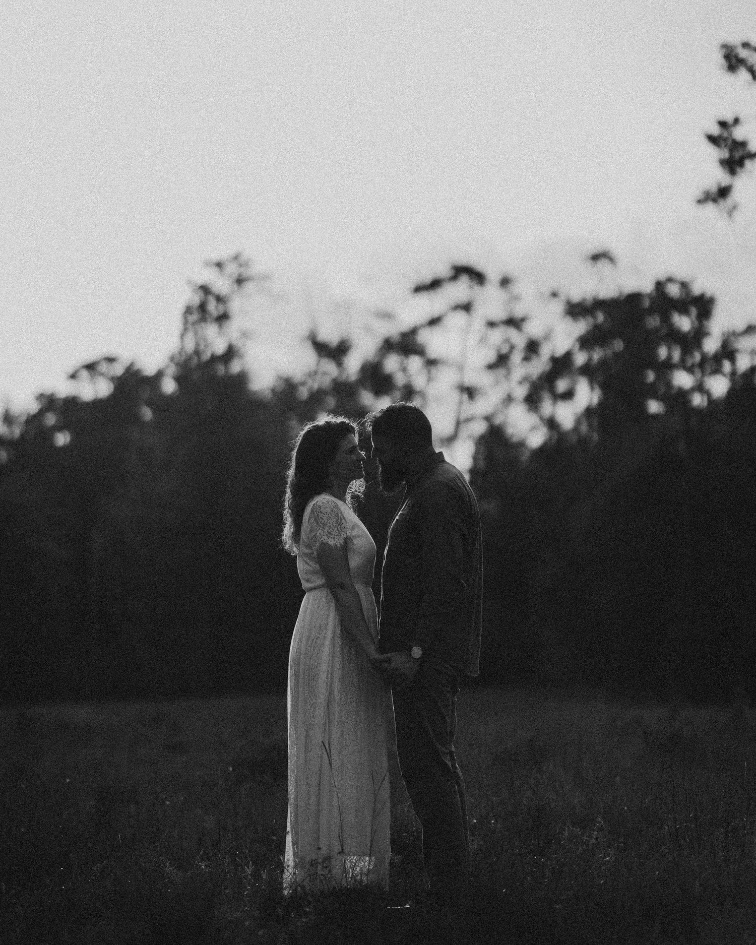 Black and white photo of a couple holding hands in a field at dusk, surrounded by trees.