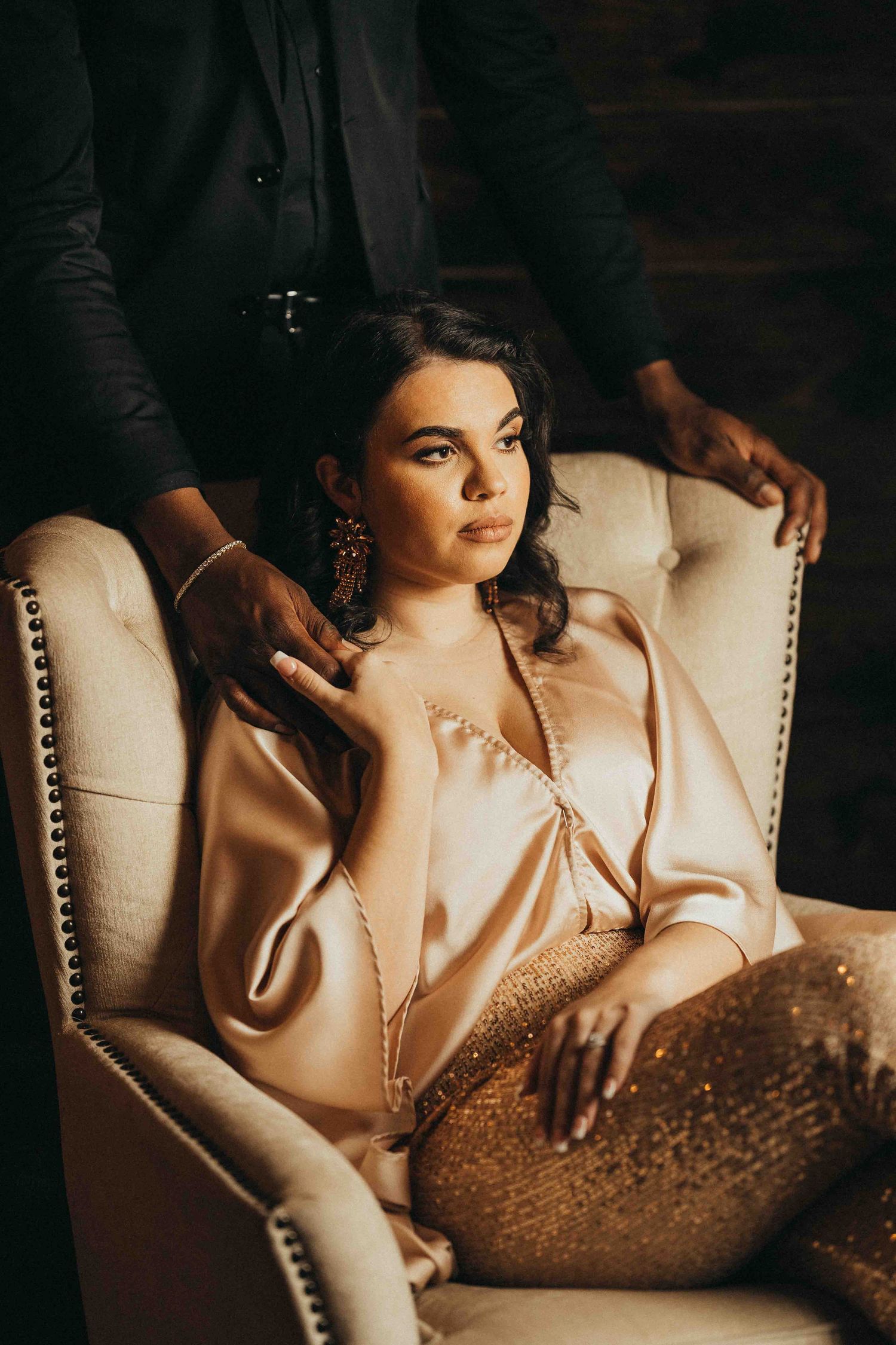Elegant woman in satin blouse and sequin pants seated in tufted chair with man standing behind her.