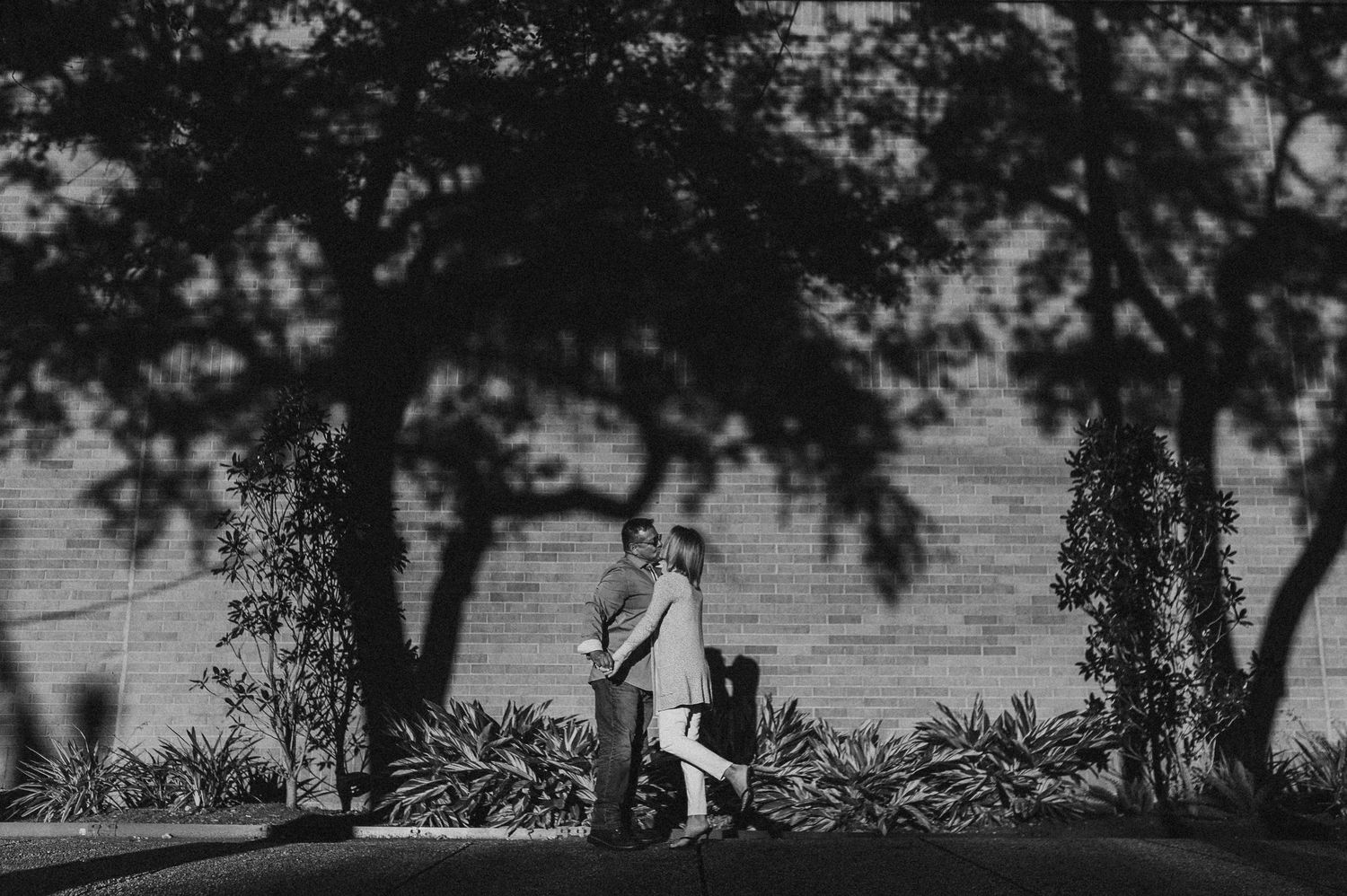 Black and white photo of a couple embracing under trees with shadows on a brick wall backdrop.