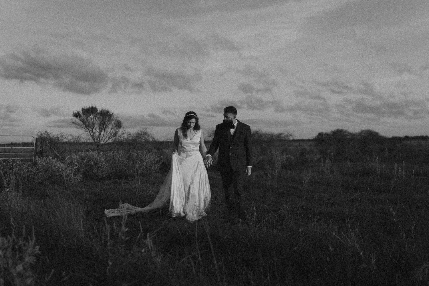 Black and white photo of a bride and groom holding hands walking through a field at dusk.