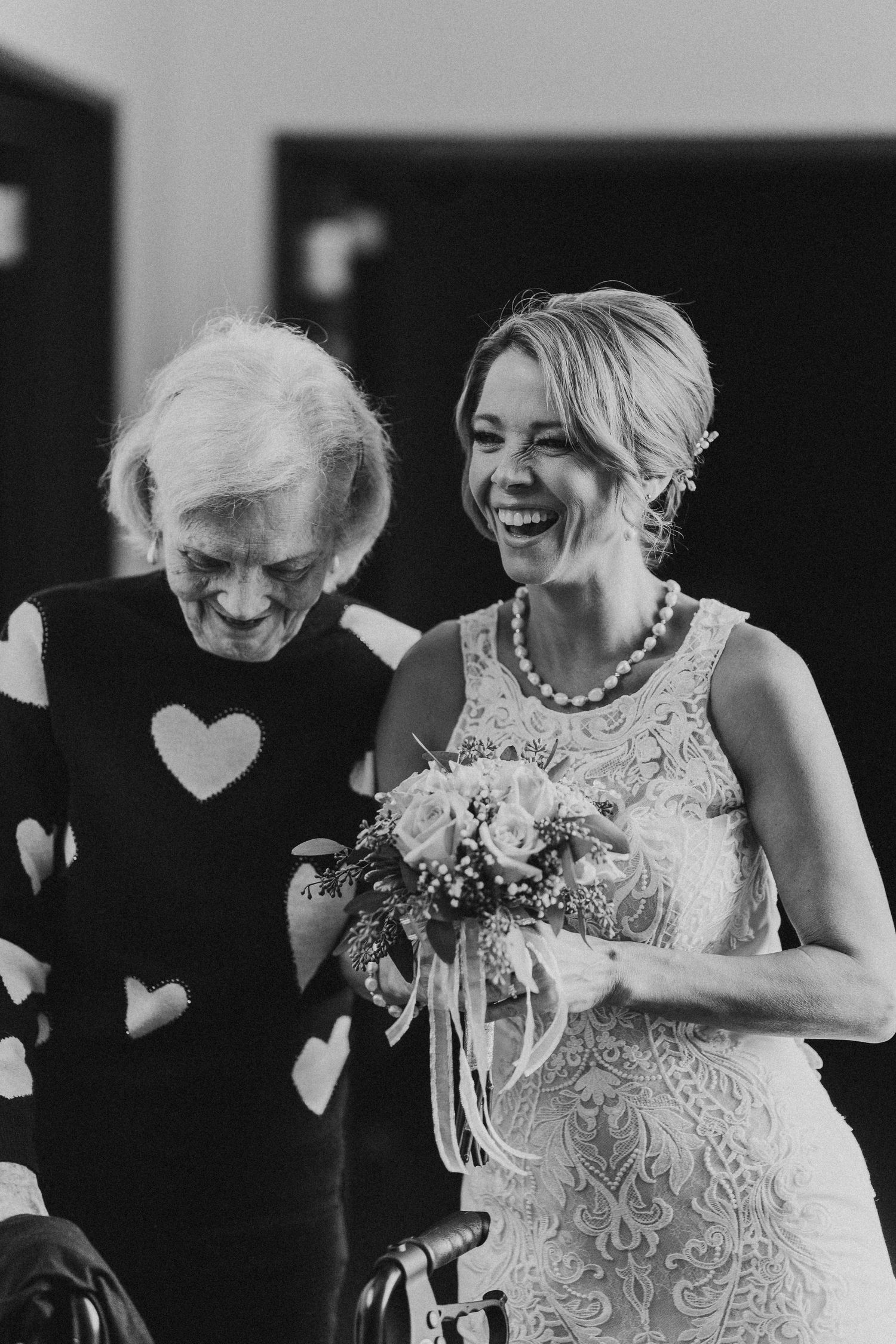 Bride in lace gown laughing joyfully with elderly woman in black and white photo.