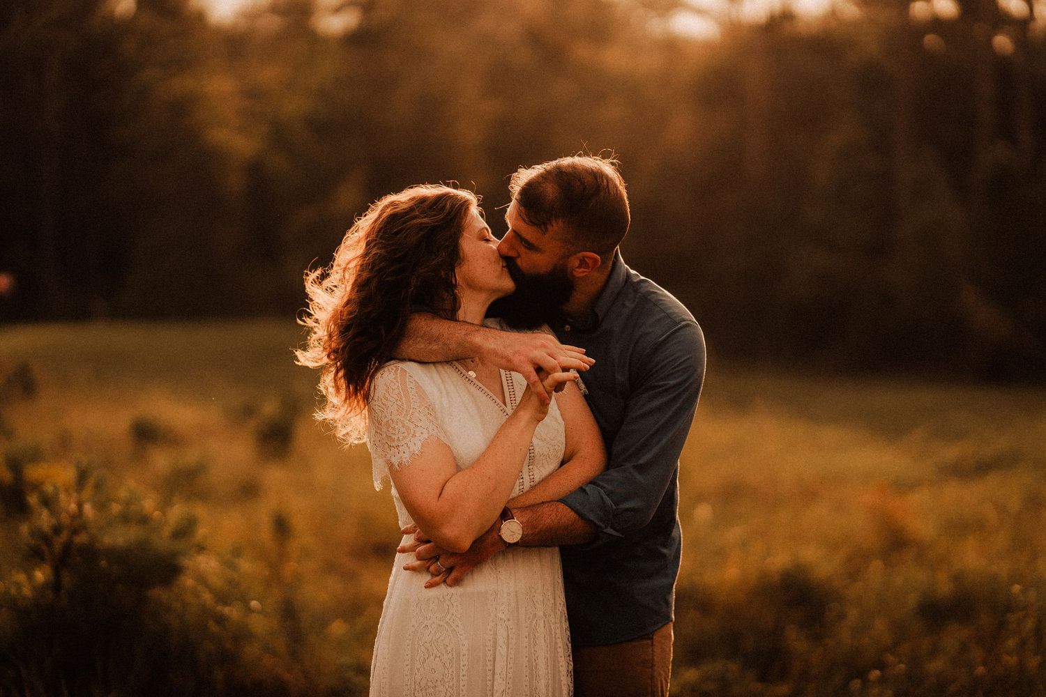 Couple embracing and kissing at golden hour in a sunlit meadow, man in blue shirt, woman in white dress.