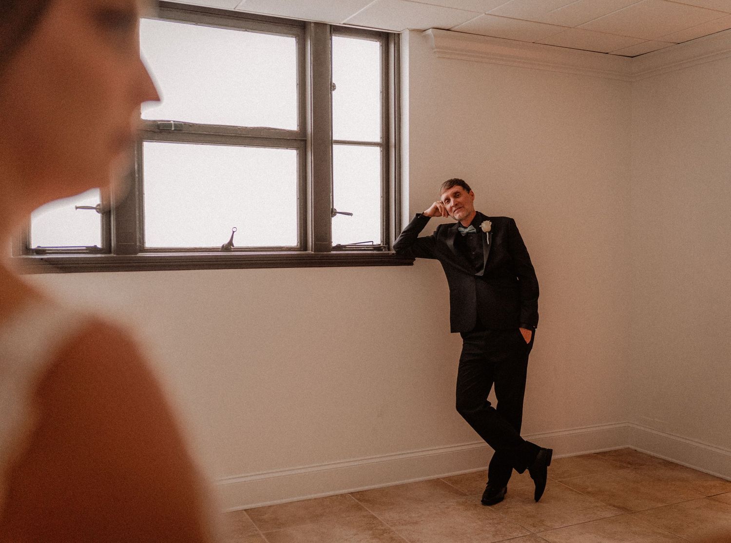 Groom in black suit leans against wall smiling at bride in bright windowed room during wedding day.