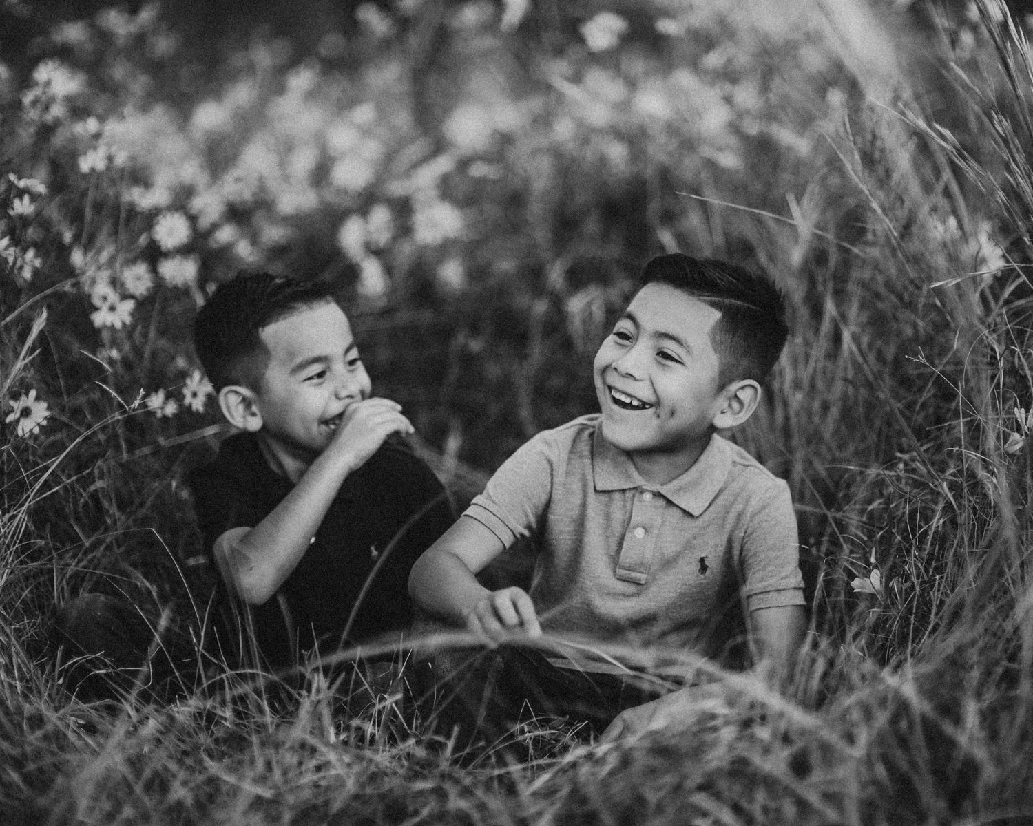Two young boys laughing and playing together in tall grass and wildflowers in a black and white photo.