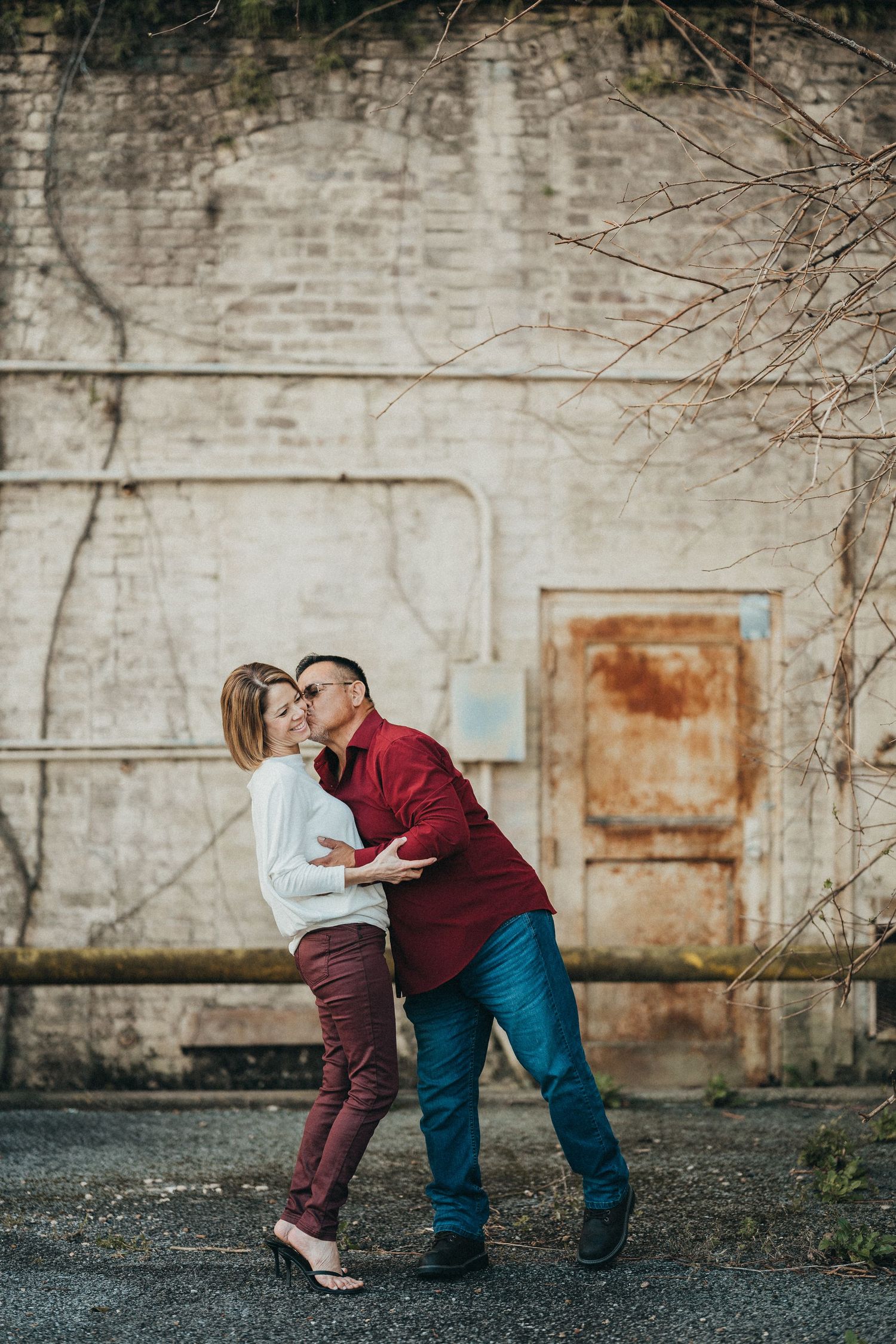 Couple sharing a romantic kiss against a rustic brick wall, man in red shirt dipping woman in burgundy pants outdoors.