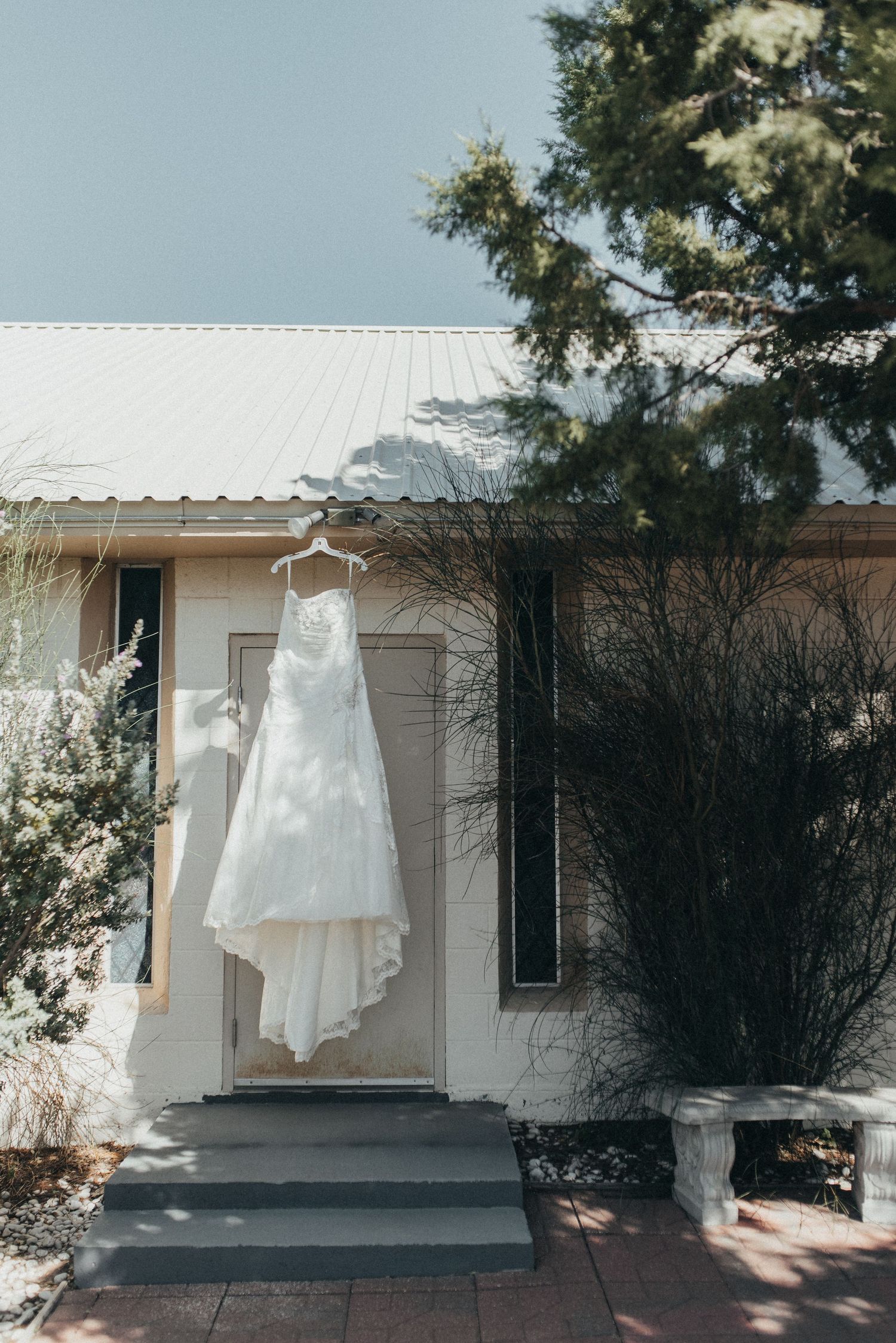 A white wedding dress hangs on a hanger outside a rustic white building surrounded by green trees on a sunny day.