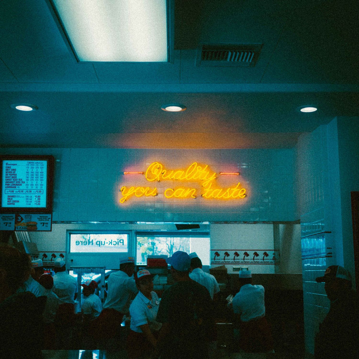 Neon sign reading Quality You Can Taste glows above a busy fast food restaurant counter filled with customers.