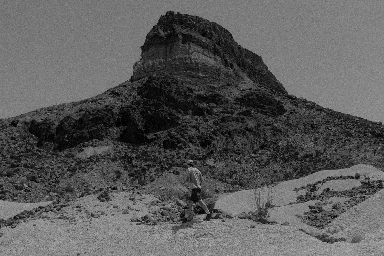 Hiker trekking through arid desert landscape with dramatic rocky butte in black and white.