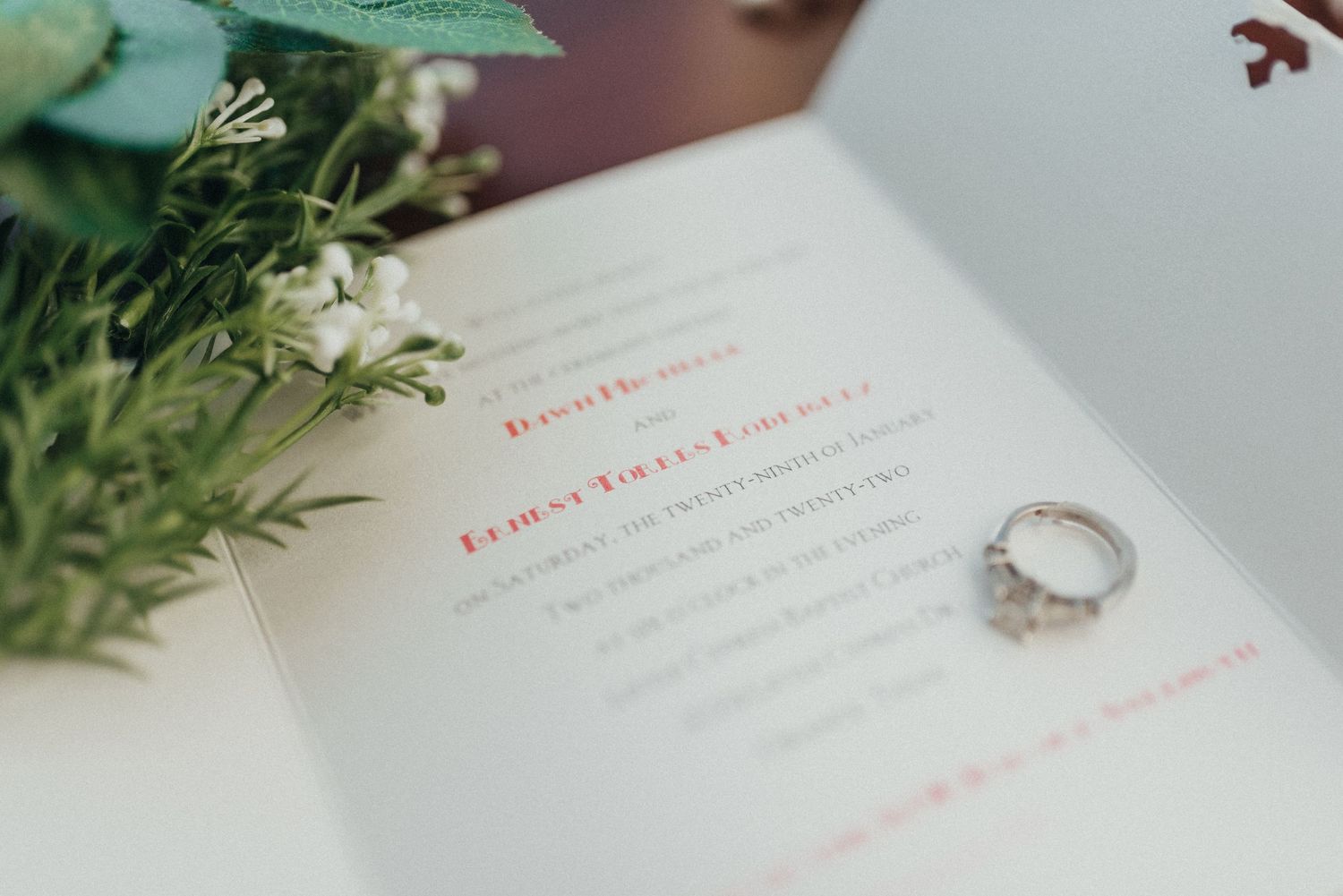 Wedding invitation with diamond engagement ring and green floral accents on white background.