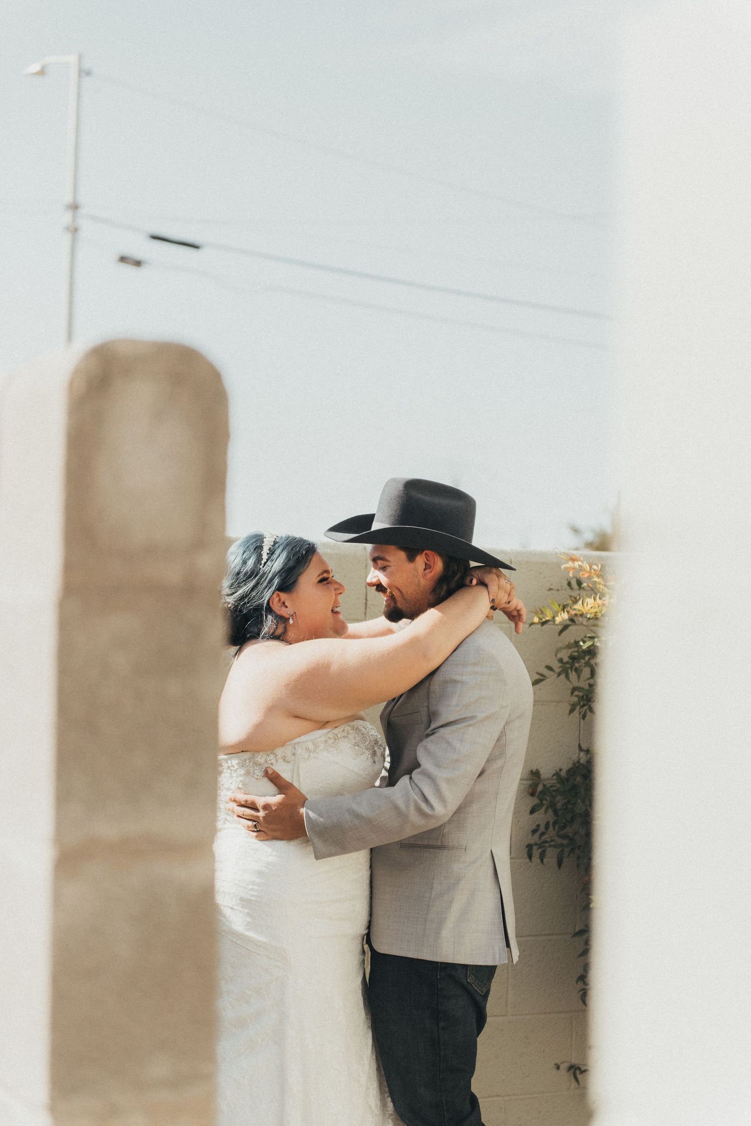 A bride and groom share a romantic embrace outdoors, with the groom wearing a black cowboy hat and gray suit.