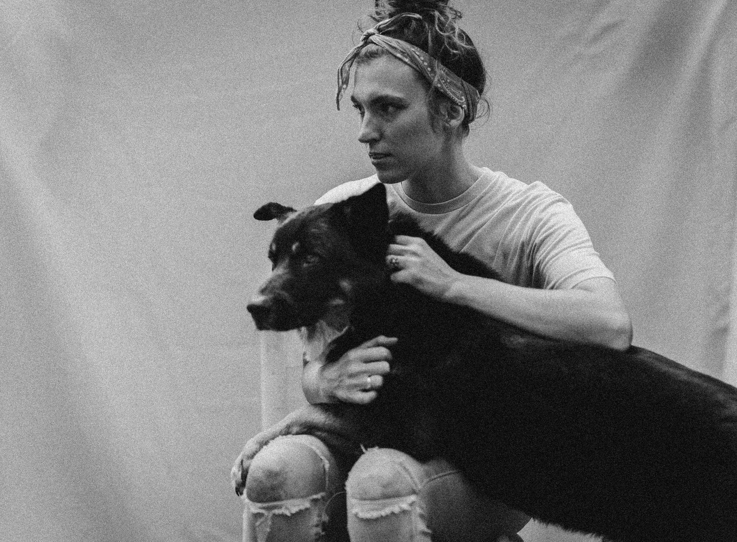 Black and white photo of a person in ripped jeans and bandana holding a large dark dog against a white backdrop.