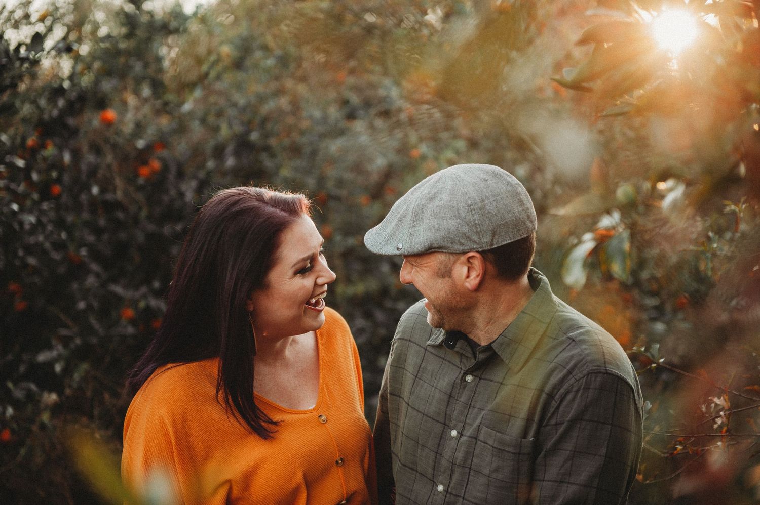 Couple smiling at each other in golden sunlight among orange trees, woman in orange top, man in grey flat cap.