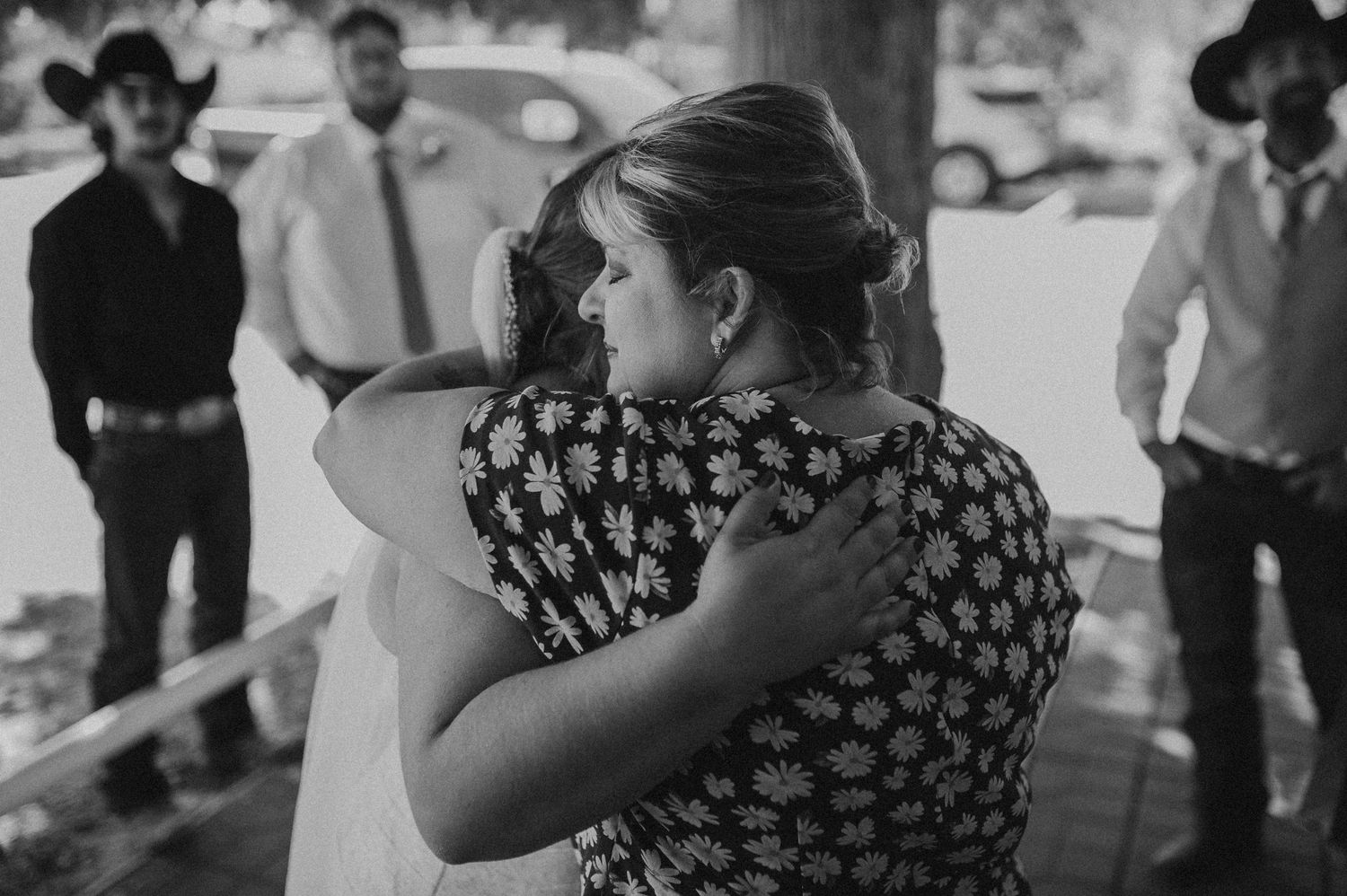 Black and white wedding photo of two women embracing warmly, with guests in cowboy hats visible in background.