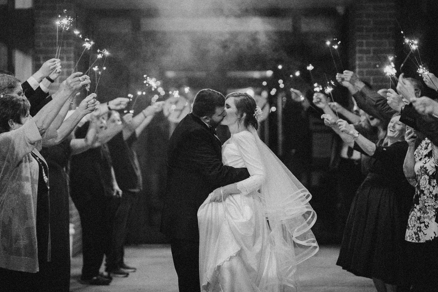 Black and white wedding photo of couple kissing under sparklers held by guests lining the exit path.