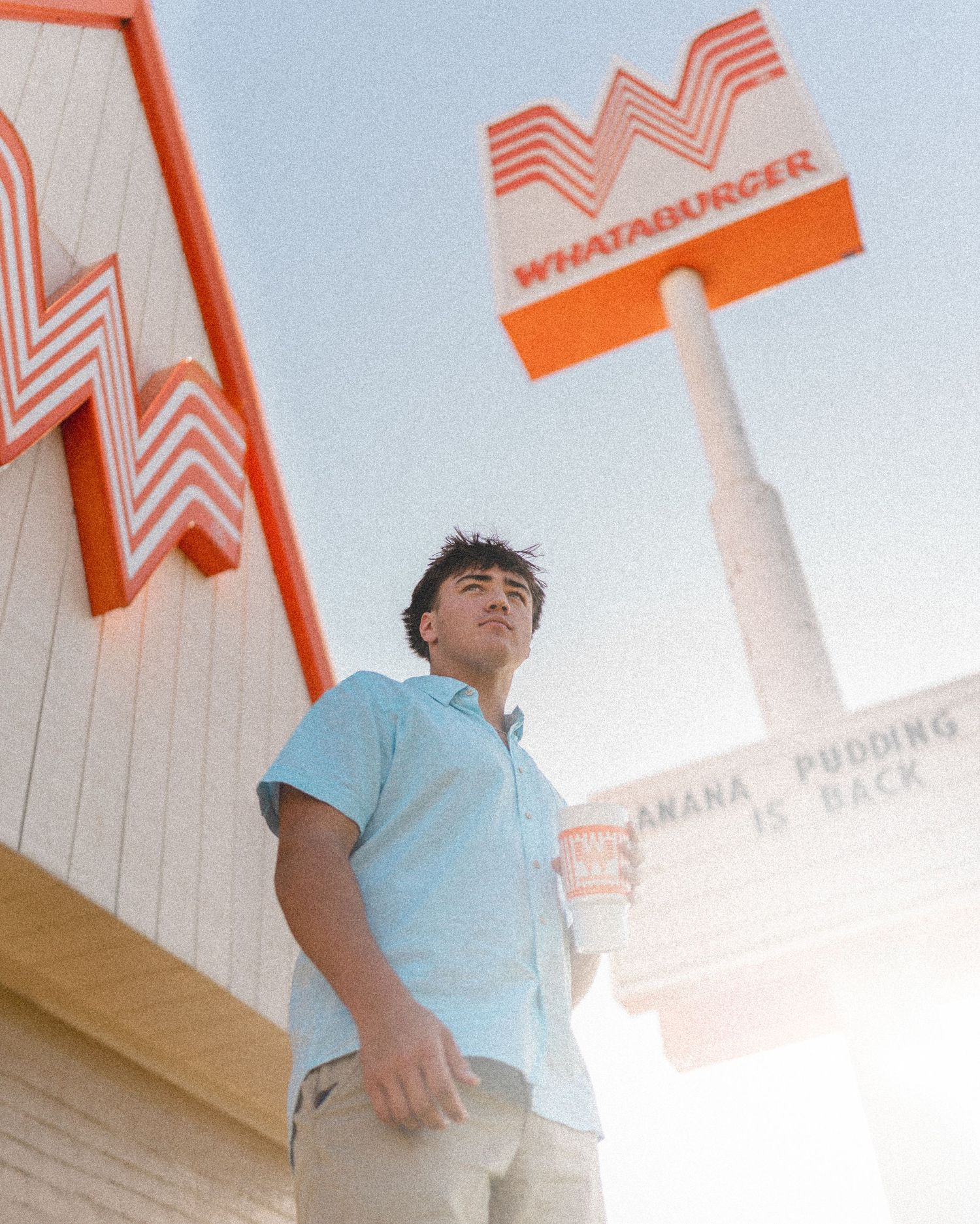 Young man in light blue shirt stands confidently beneath a Whataburger sign on a sunny day.