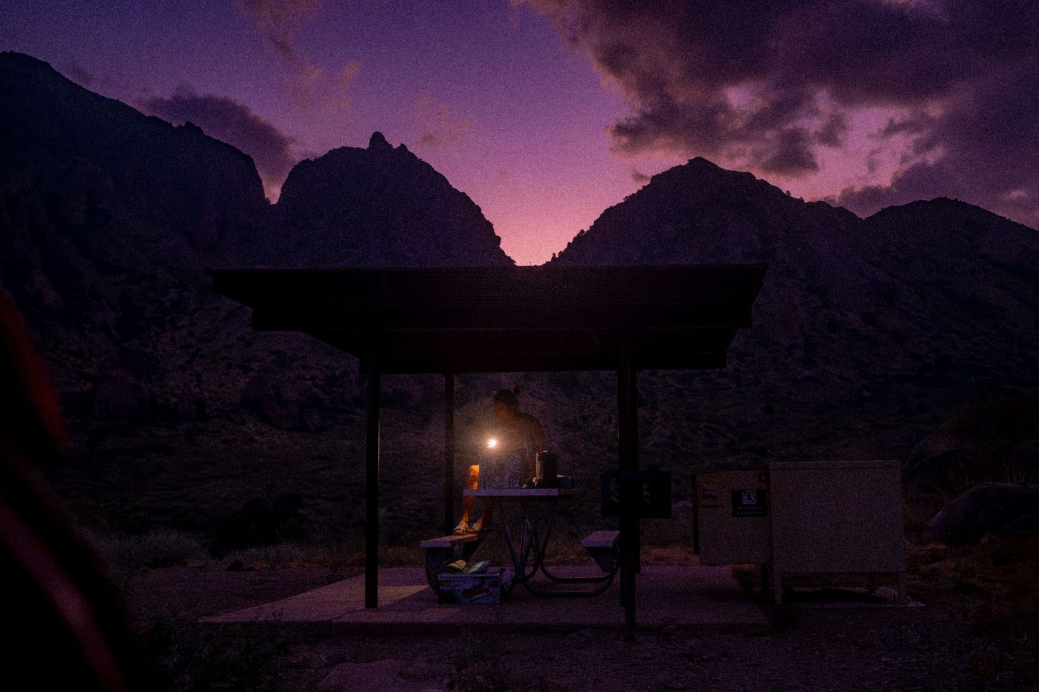 A glowing lantern illuminates a covered outdoor shelter at dusk with dramatic purple mountains in the background.