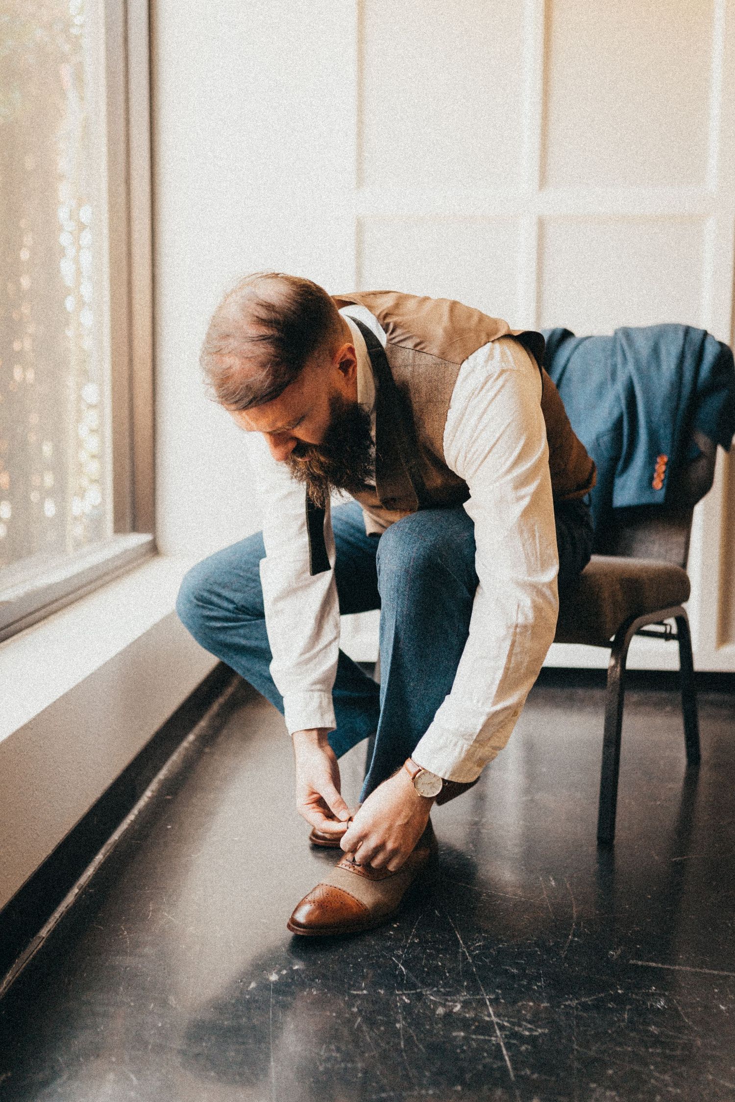 Groom tying brown leather dress shoes while seated by window before wedding ceremony.